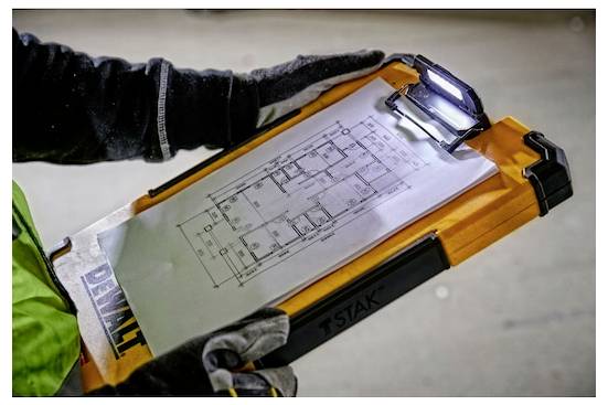 Eine Person mit Handschuhen hält ein Klemmbrett mit einem auf einer Klemme befestigten Grundrissplan. Das Klemmbrett hat oben ein Licht.