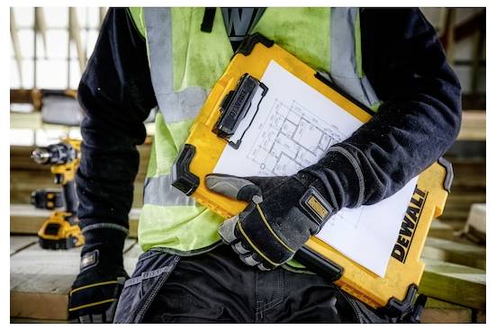 „Person in Arbeitskleidung hält ein gelbes DeWalt-Klemmbrett mit Bauplänen und steht auf einer Baustelle mit Werkzeugen im Hintergrund."