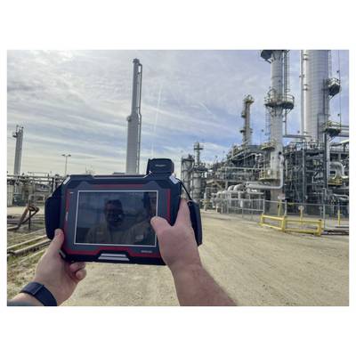 Eine Person hält ein Tablet vor einer Industrieraffinerie und erfasst möglicherweise Daten oder nutzt es als Anleitung für Inspektionszwecke.