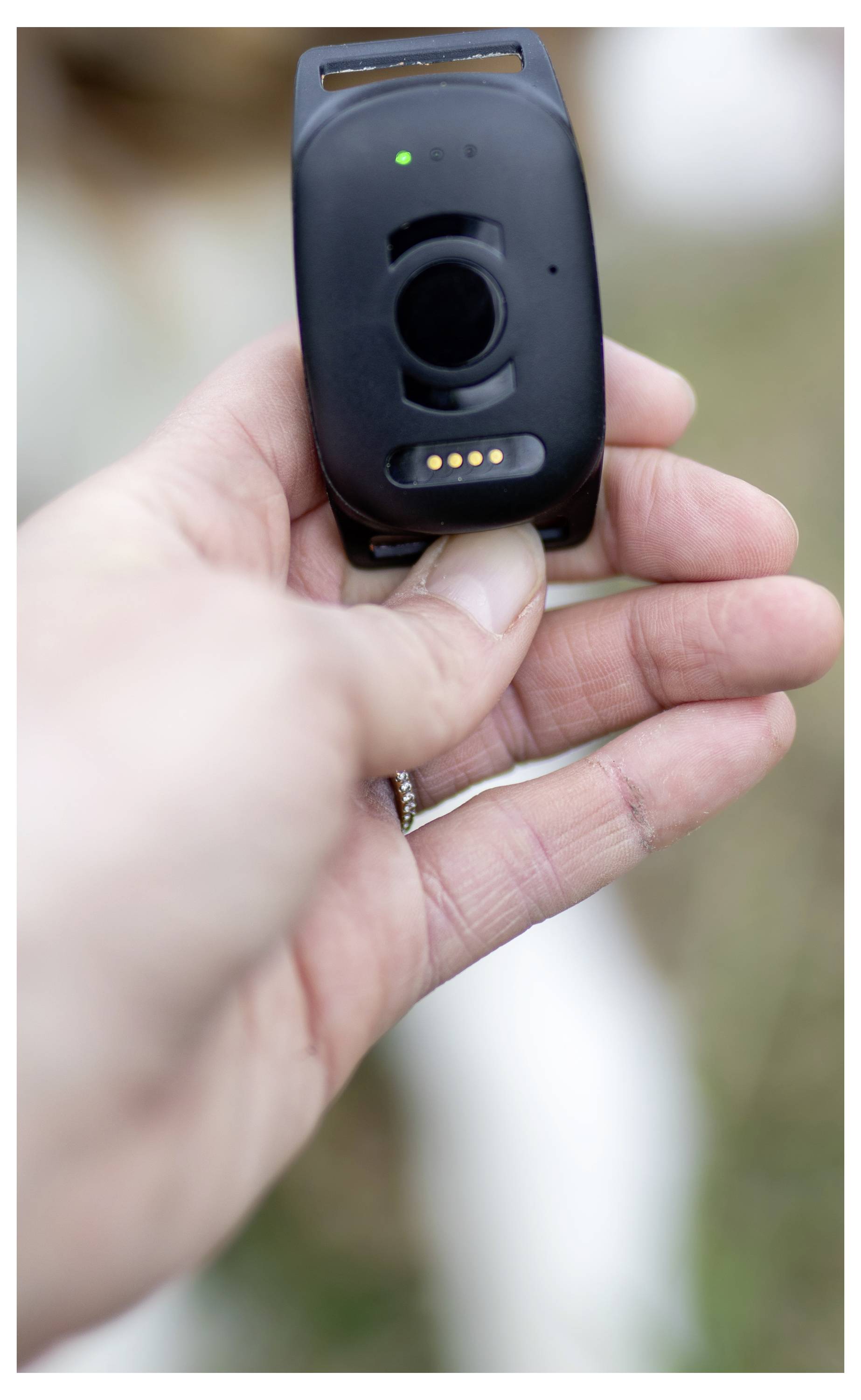 Eine Hand, die ein schwarzes GPS-Ortungsgerät im Freien hält, möglicherweise für ein Haustier oder eine Person, mit einer grünen Statusleuchte, die anzeigt, dass es aktiv ist.