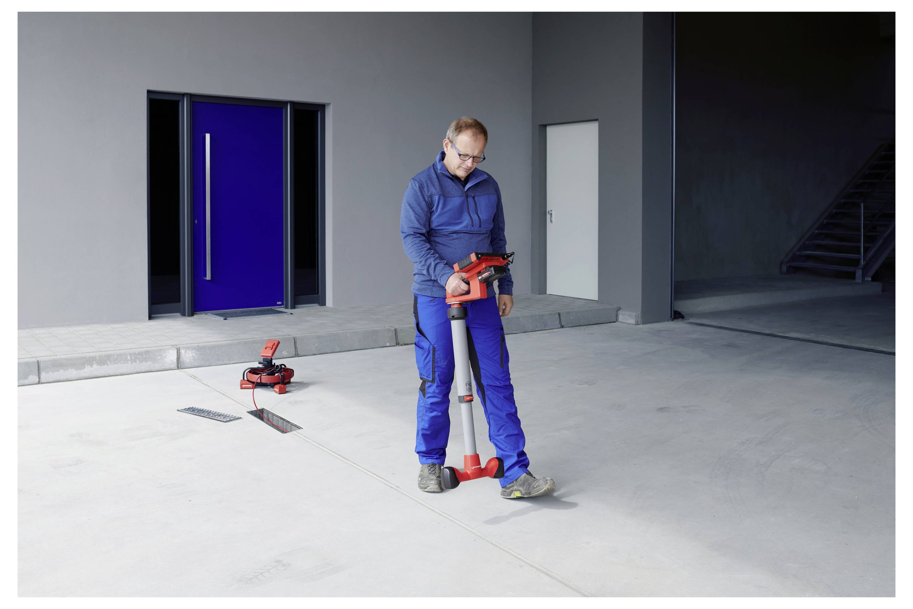 Eine Person in blauer Kleidung verwendet ein rotes und graues Bodenmesswerkzeug in einer geräumigen, industriellen Innenumgebung mit großen Türen.