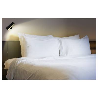 Ein sauber gemachtes Bett mit weißen Kissen und einer Bettdecke in einem sanft beleuchteten Hotelzimmer mit einer an der Wand angebrachten Leselampe über dem Kopfende.