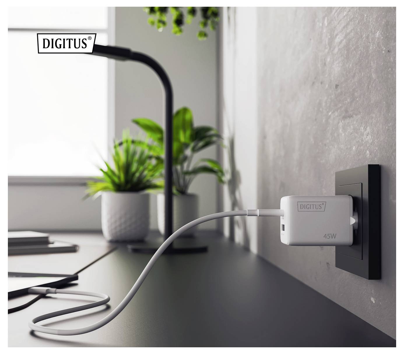 Ein weißes 45-W-Ladegerät von DIGITUS, das in eine Wandsteckdose auf einem schwarzen Schreibtisch eingesteckt ist. Eine Lampe und eine Topfpflanze befinden sich im Hintergrund.