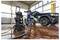 Eine Person in blauem Arbeitsanzug verwendet einen Hochdruckreiniger, um ein schwarzes Auto in einer hellen Garage mit Fliesenboden und großen Fenstern zu reinigen.