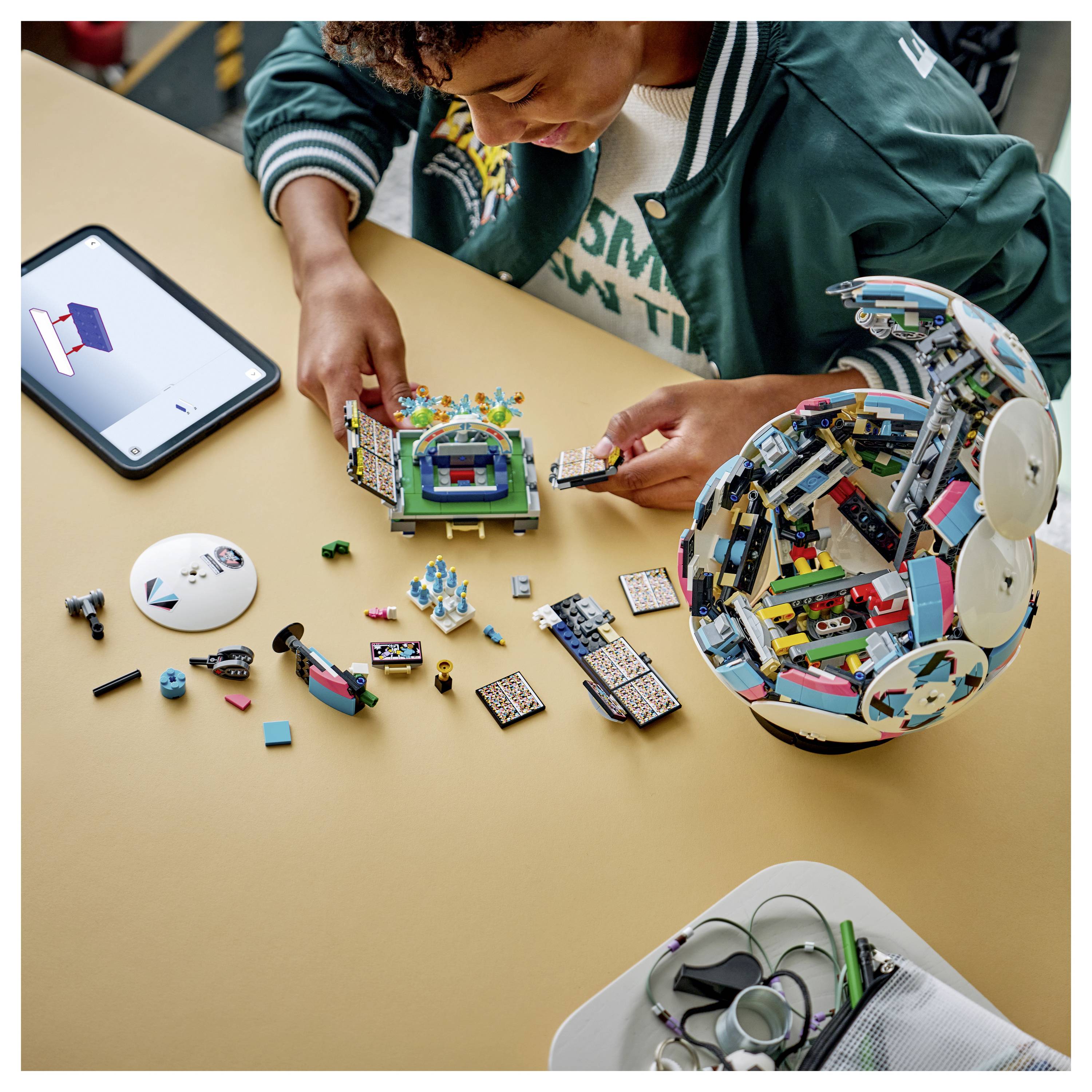 Eine Person baut ein farbiges LEGO-Set auf einem Tisch zusammen. Ein Tablet zeigt Anweisungen an, und verschiedene kleine LEGO-Steine sind rundherum verstreut.
