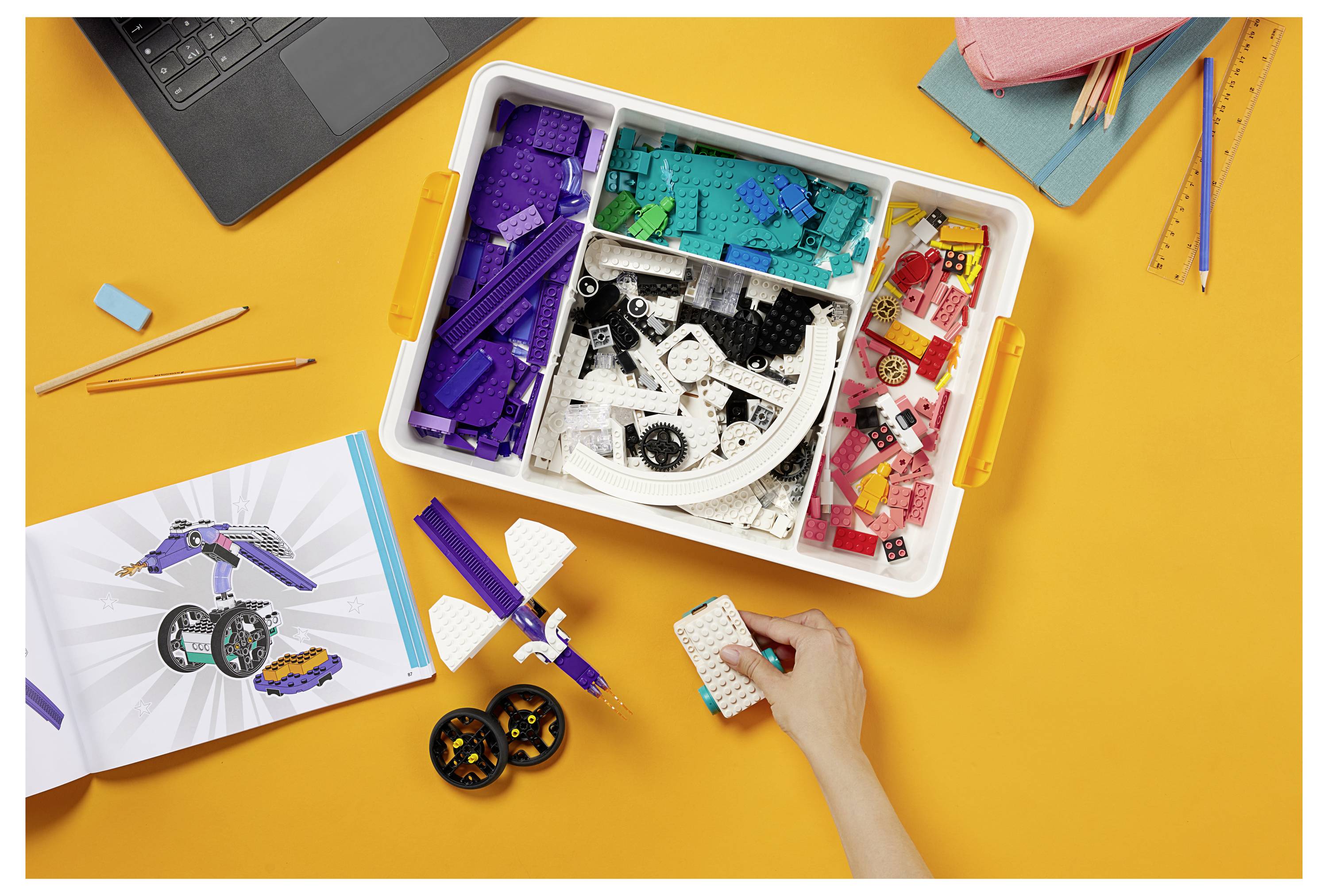 Eine Person setzt ein Projekt aus LEGO-Steinen eines Sets zusammen, wobei eine Anleitung und ein Laptop in der Nähe auf einer leuchtend orangefarbenen Oberfläche liegen.