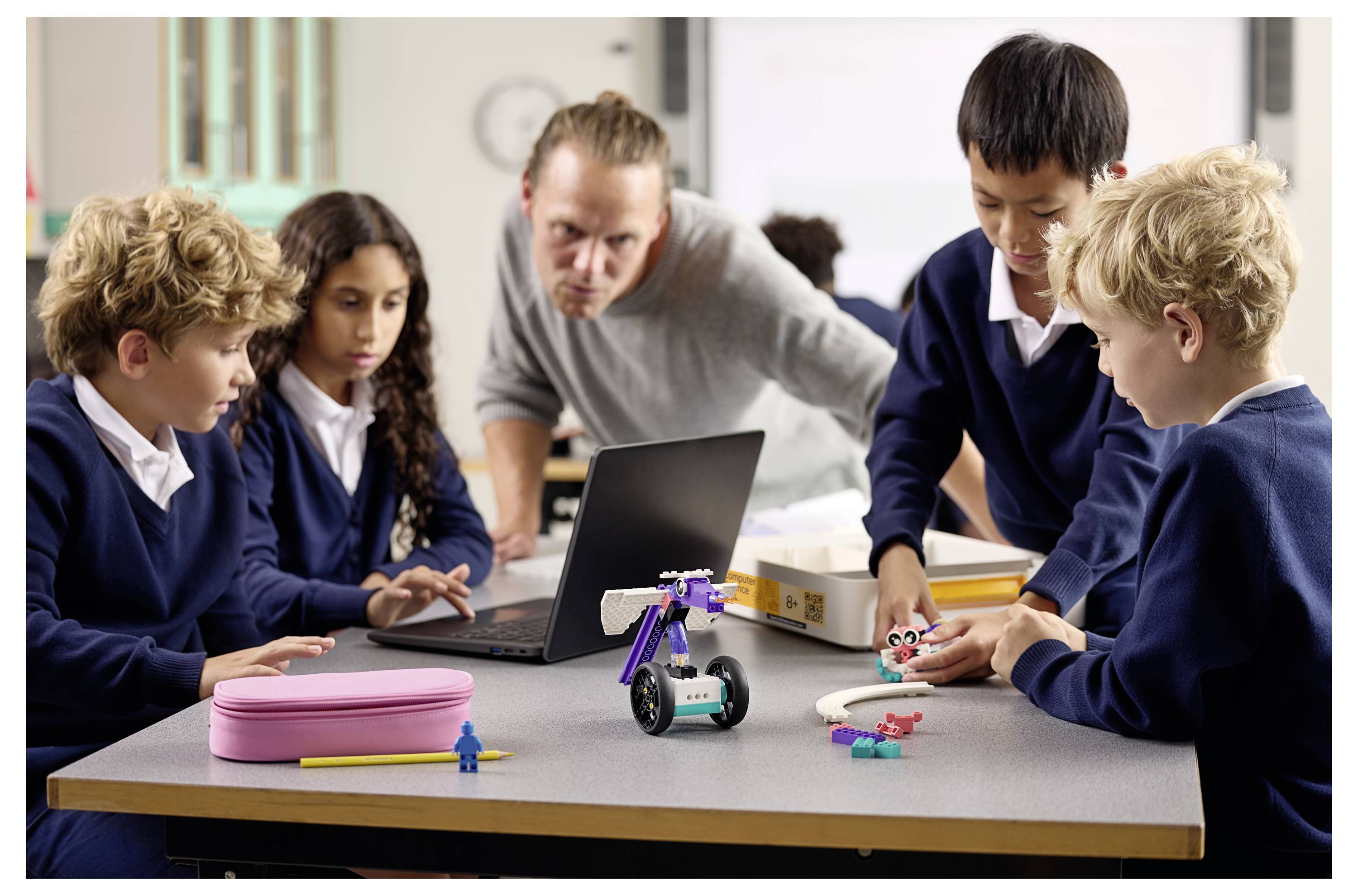 Eine Gruppe von Schülern und ein Lehrer bauen mit Bausätzen und einem Laptop im Klassenzimmer Roboter und arbeiten an einem MINT-Projekt zusammen.