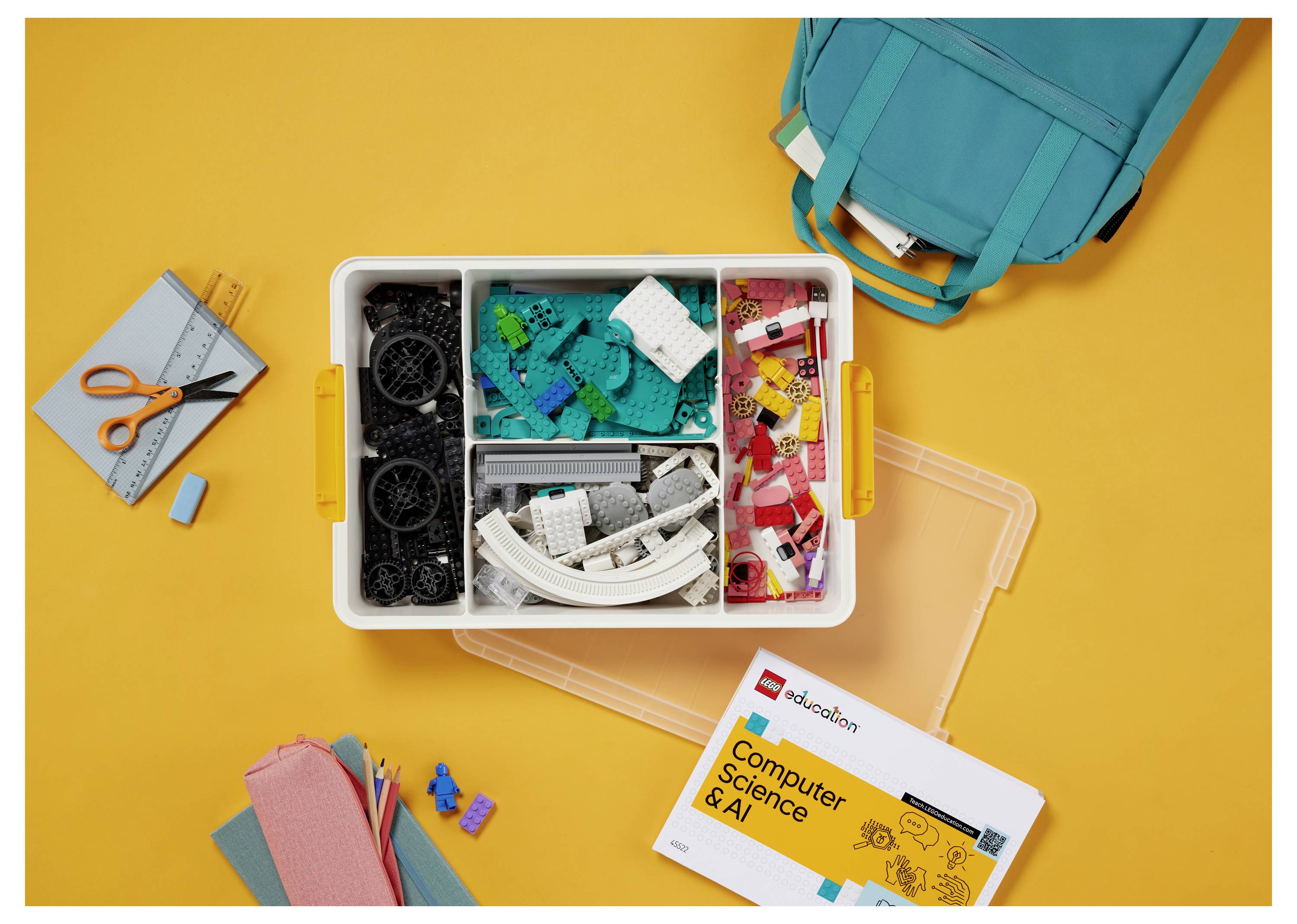 Eine Kiste mit nach Farben sortierten Legosteinen steht auf einer orangefarbenen Oberfläche. In der Nähe befinden sich ein blaugrüner Rucksack, ein Notizbuch, ein Stiftemäppchen und eine KI-Broschüre.