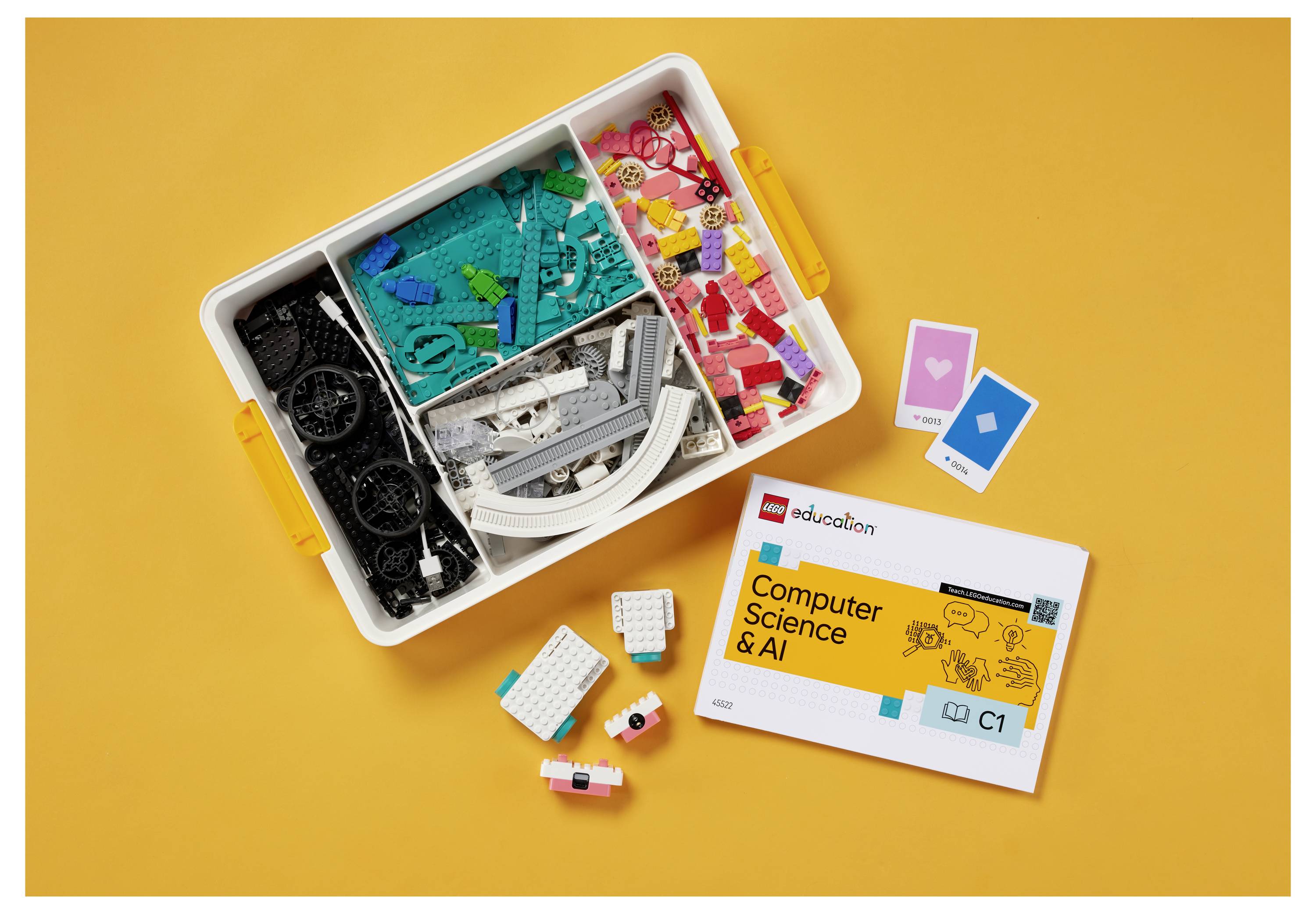 Eine Schachtel mit bunten LEGO-Steinen ist geöffnet, zusammen mit Heften mit dem Titel „Computer Science & AI", vor einem orangefarbenen Hintergrund.