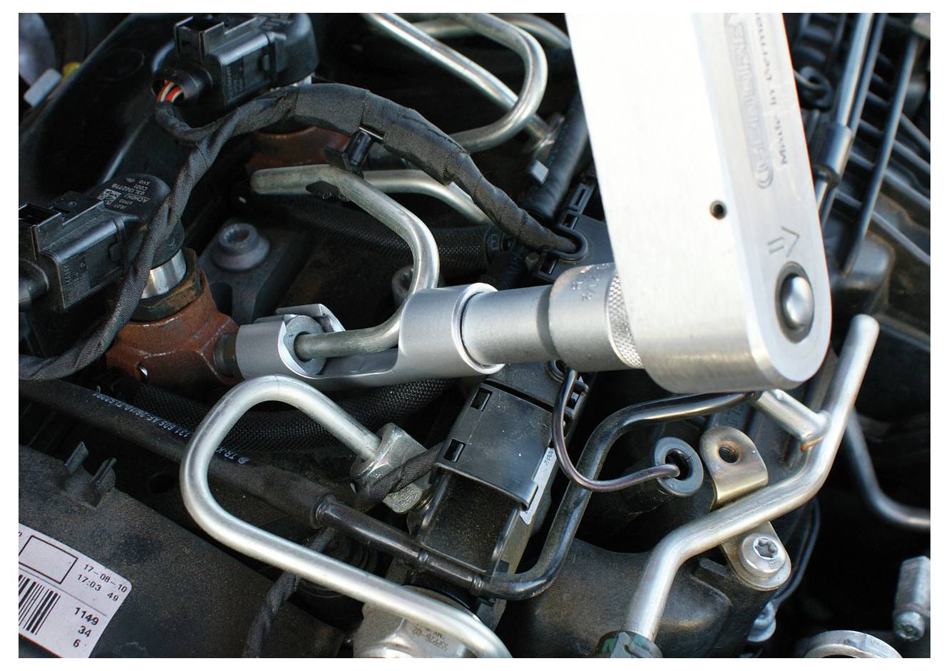 Nahaufnahme eines Drehmomentschlüssels, der eine Schraube an einem Motorenbauteil anzieht, umgeben von Metallrohren und Verkabelung in einem Motorraum eines Autos.