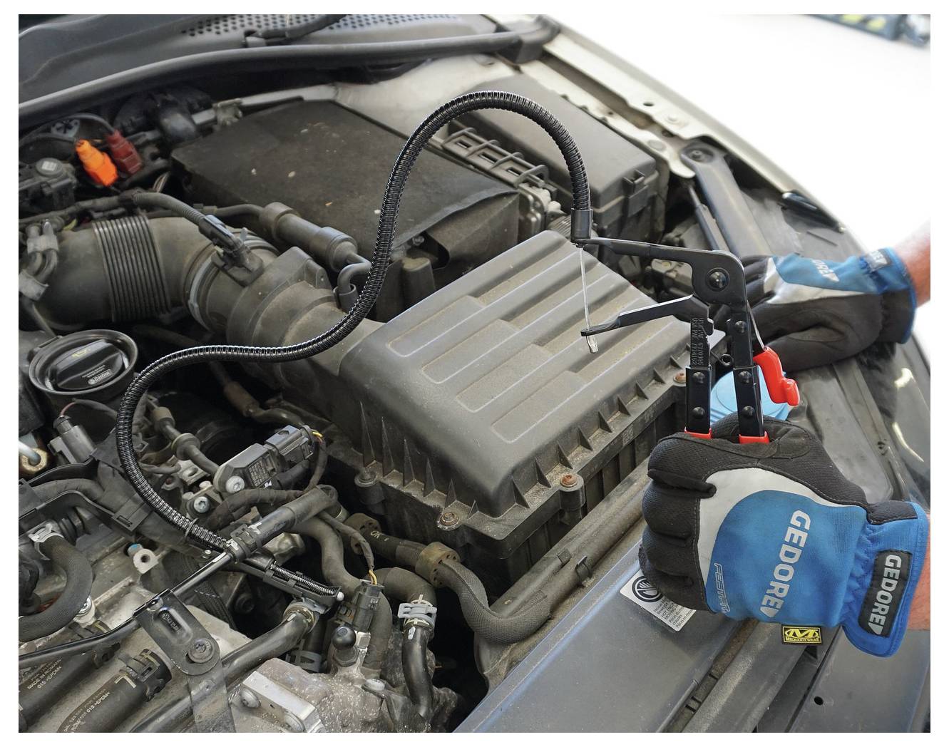 „Hände, die ein Werkzeug zur Überprüfung des Automotors verwenden; Nahaufnahme von Motorkomponenten und Werkzeug mit flexiblem Kabel. Handschuhe zeigen die Marke „Gedore"."