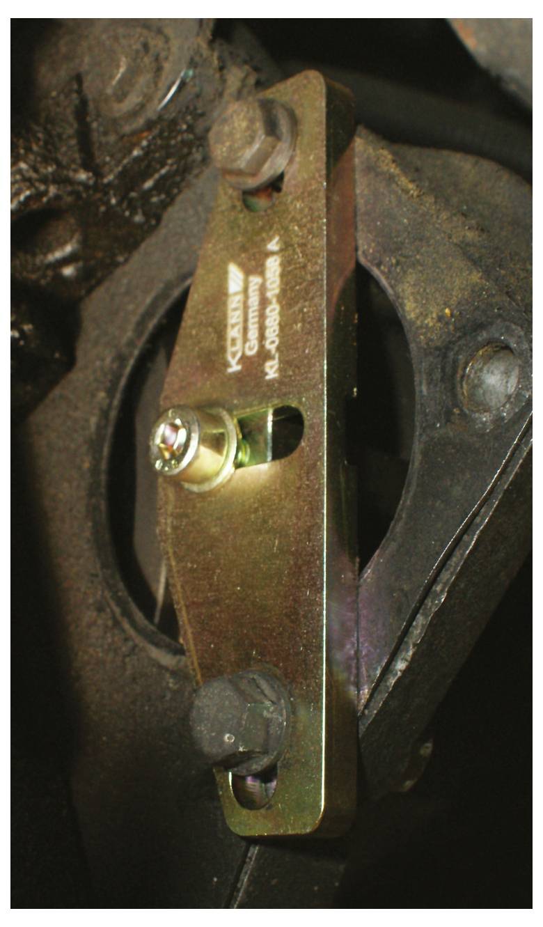 Ein Metallteil für Kraftfahrzeuge ist mit drei Bolzen an einer dunklen Maschinenkomponente montiert. Der Text auf dem Teil lautet „Caster, Camber".