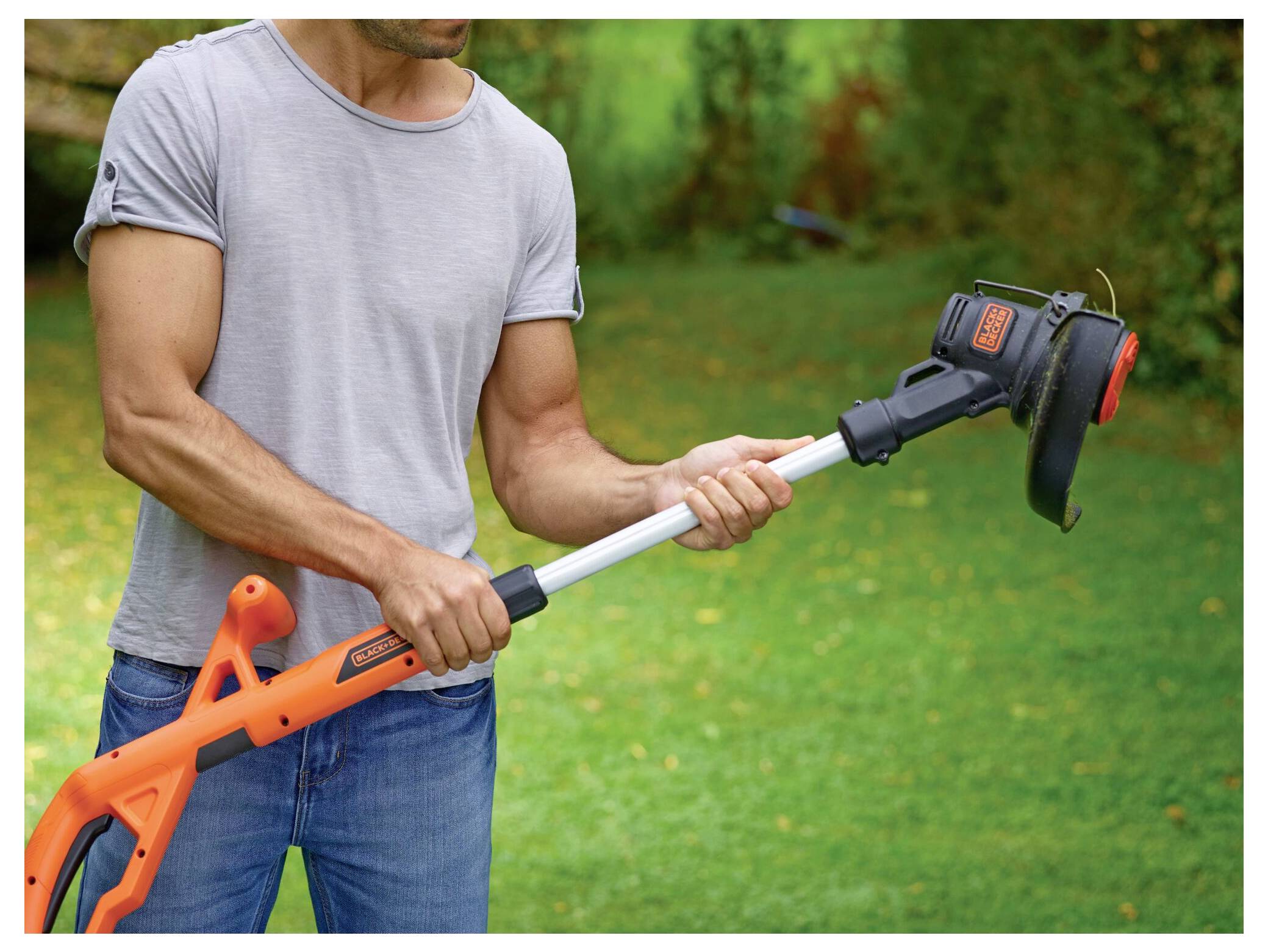 Eine Person, die einen orange-schwarzen Rasentrimmer hält und einstellt, mit einem Garten im Hintergrund, wobei der Fokus auf dem Griff und dem Kopf des Geräts liegt.