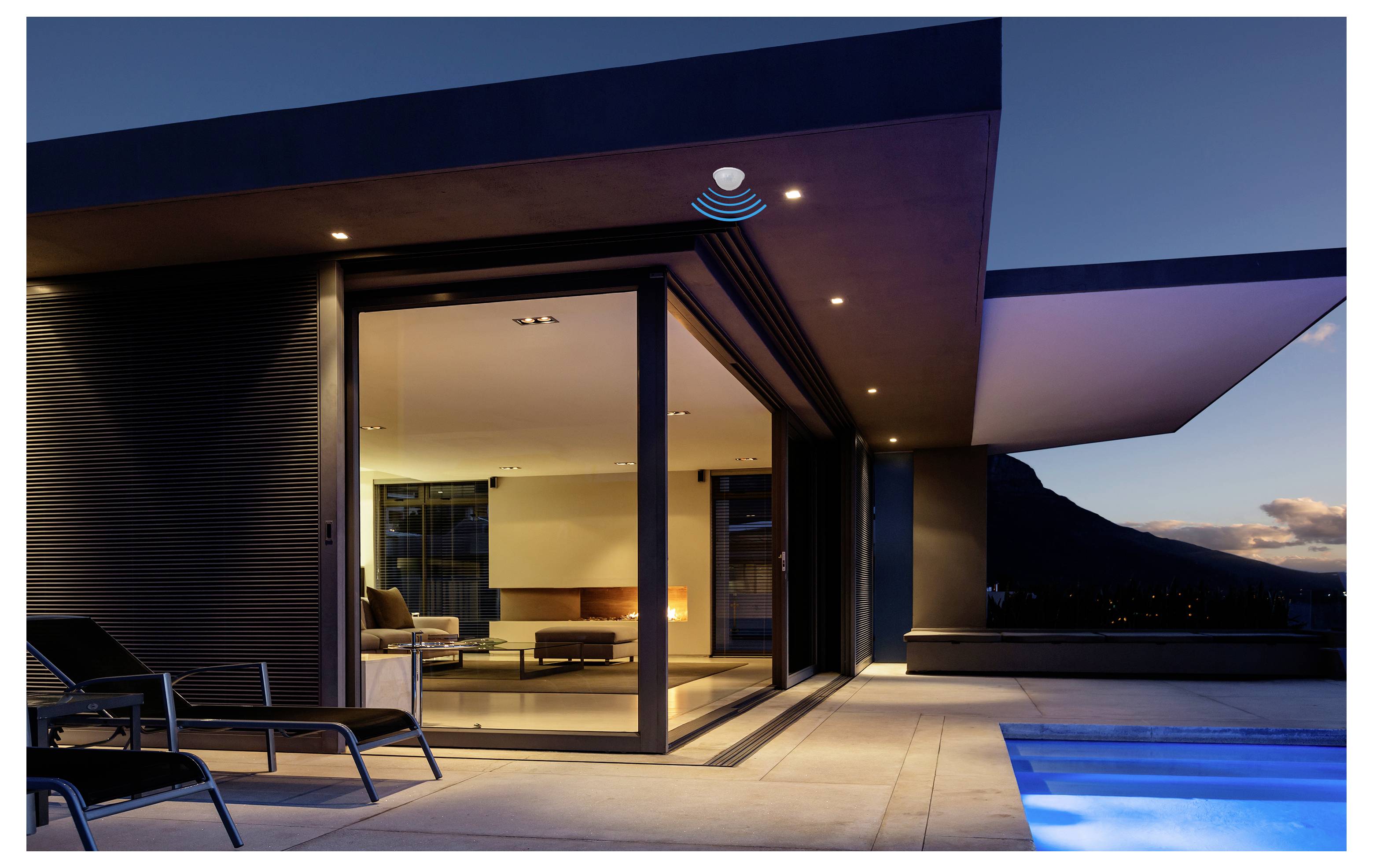 Moderne Luxus-Terrasse mit geöffneten Glasschiebetüren zum beleuchteten Wohnzimmer, Liegestühle davor und leuchtenden Pool bei Dämmerung mit Bergblick.