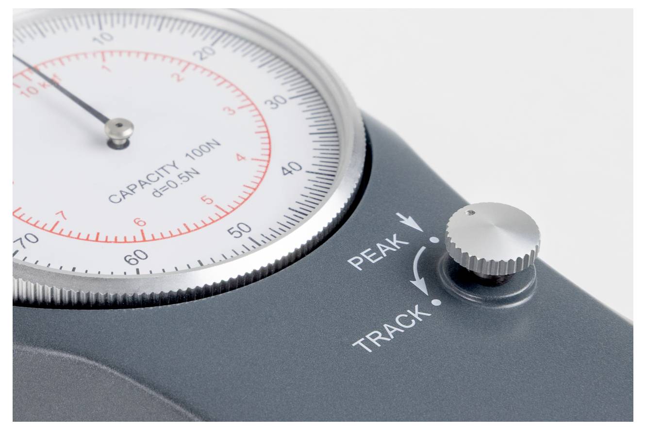 Eine Nahaufnahme eines mechanischen Messgeräts mit einer Nadel, die auf einer kreisförmigen Skala auf etwa 90 zeigt, mit der Angabe "Kapazität 100N". Ein Drehregler stellt die Einstellungen "Peak" und "Track" ein.