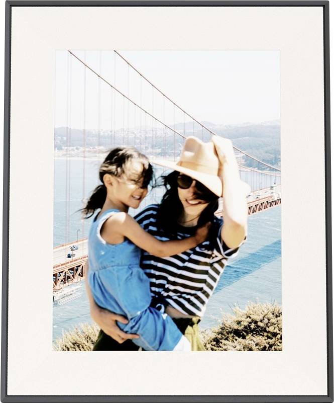 Eine Frau in einem gestreiften Hemd und Hut hält ein lachendes kleines Mädchen in einem blauen Kleid. Die Golden Gate Bridge ist im Hintergrund sichtbar.