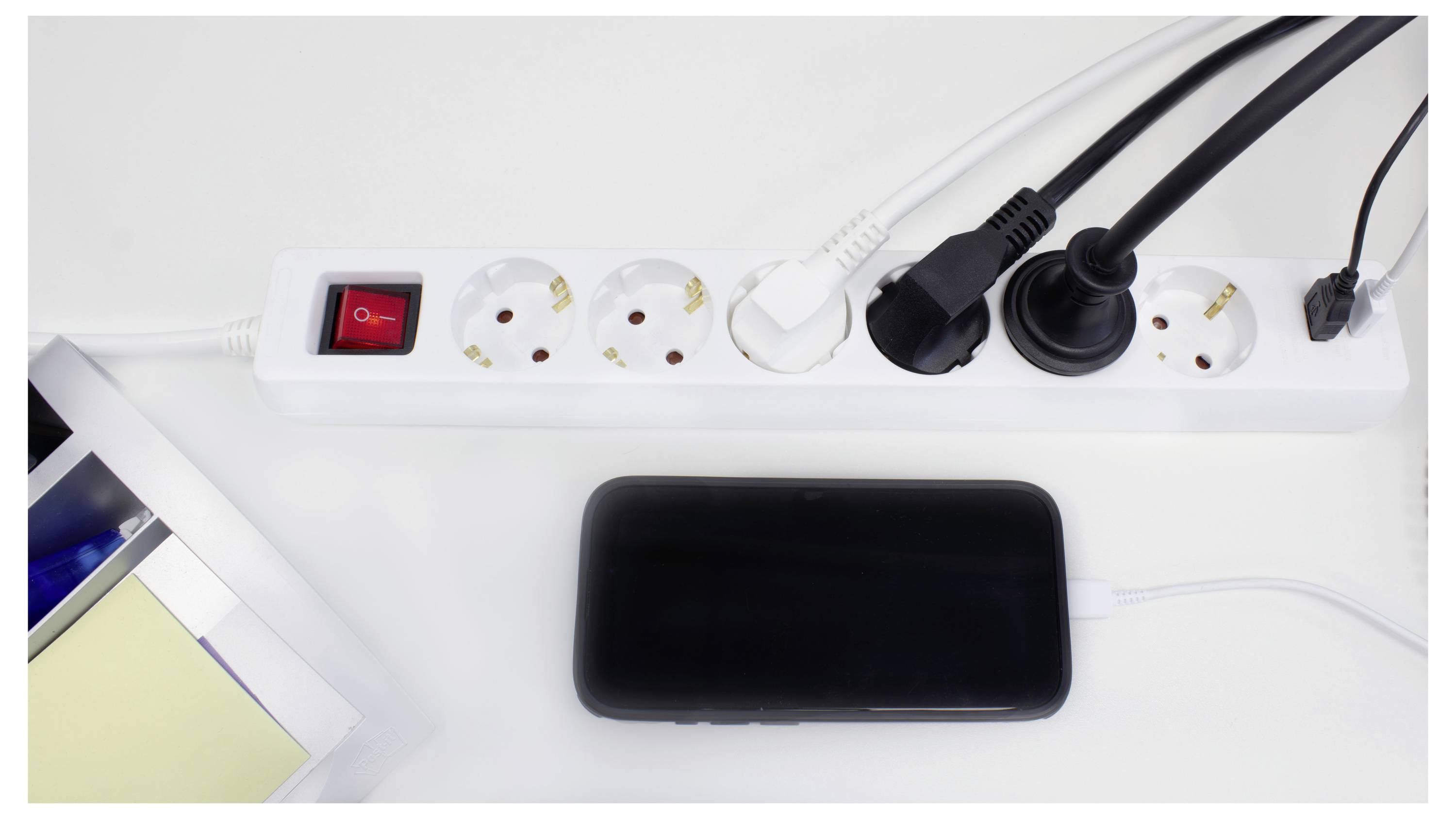 Ein weißes Steckerleistenset mit mehreren angeschlossenen Geräten, einschließlich Telefonladegeräten und einem schwarzen Smartphone, das auf einem weißen Schreibtisch angeschlossen ist.
