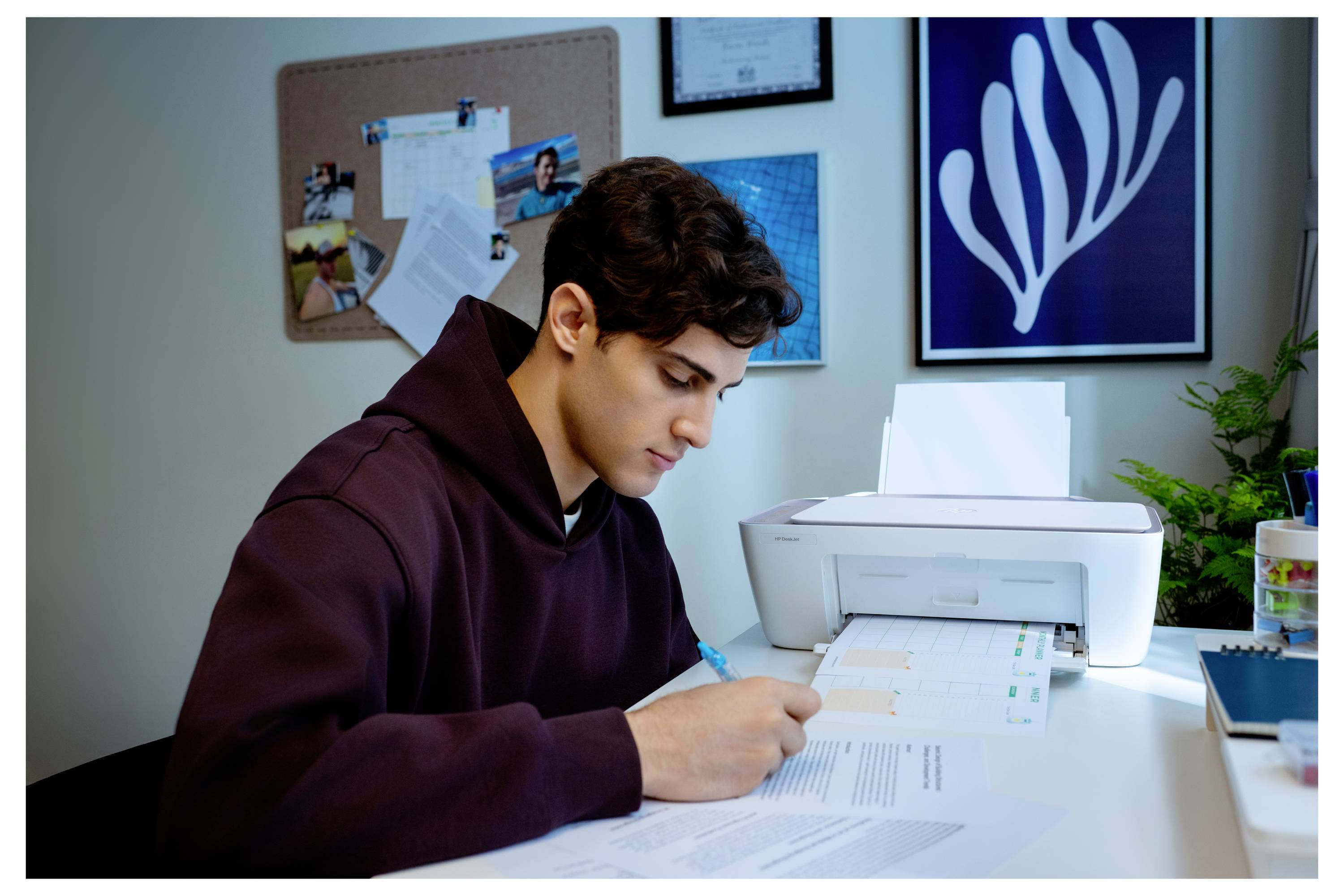 Eine Person in einem Hoodie sitzt an einem Schreibtisch und schreibt auf Papieren, während sich ein Drucker in der Nähe befindet. An der Wand sind ein Bulletin-Board und gerahmte Kunstwerke zu sehen.