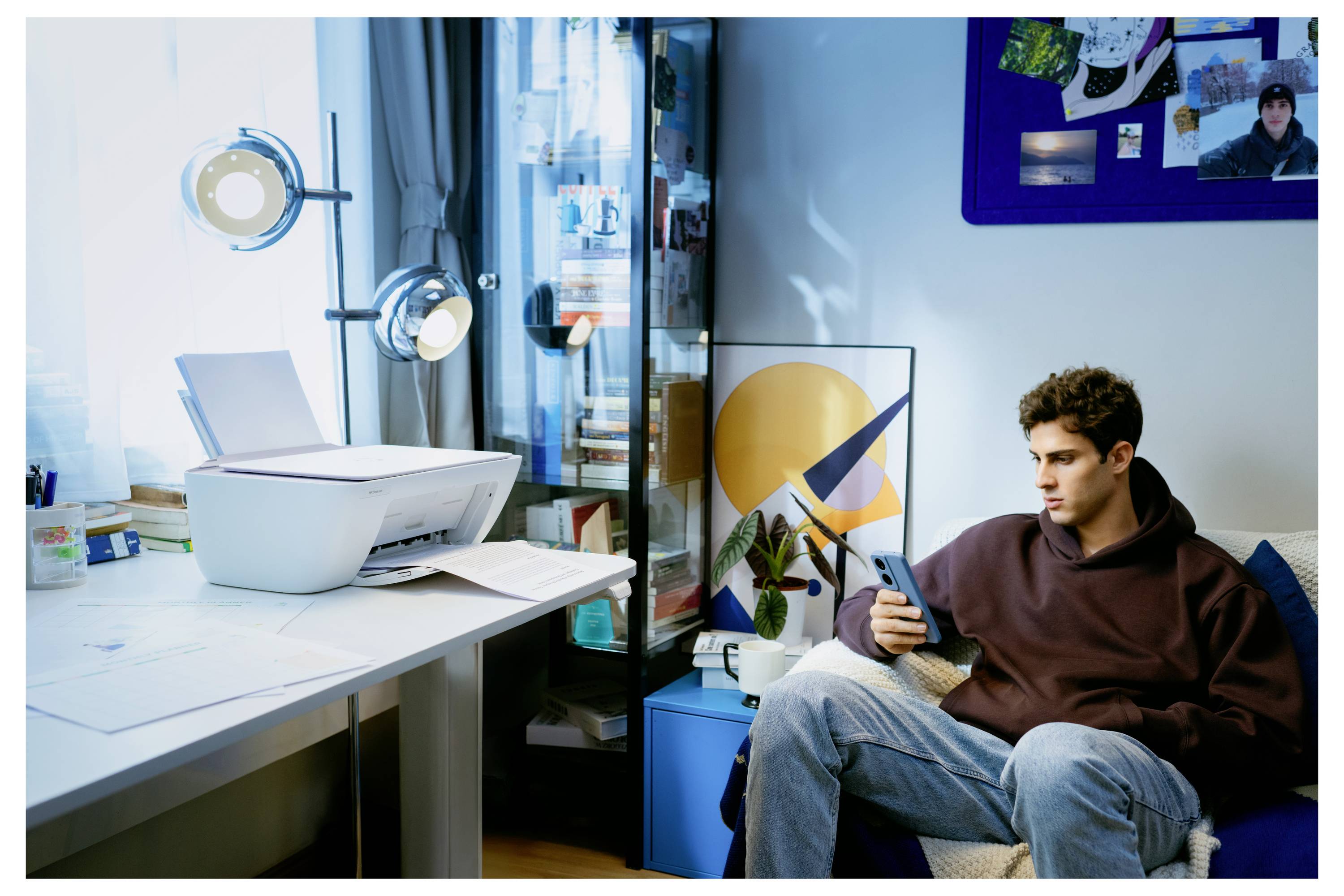 Eine Person sitzt auf einem Sofa und schaut auf ein Smartphone, neben einem Tisch mit einem Drucker und Papieren. Ein Schrank mit Büchern und Kunstdekoration ist in der Nähe.