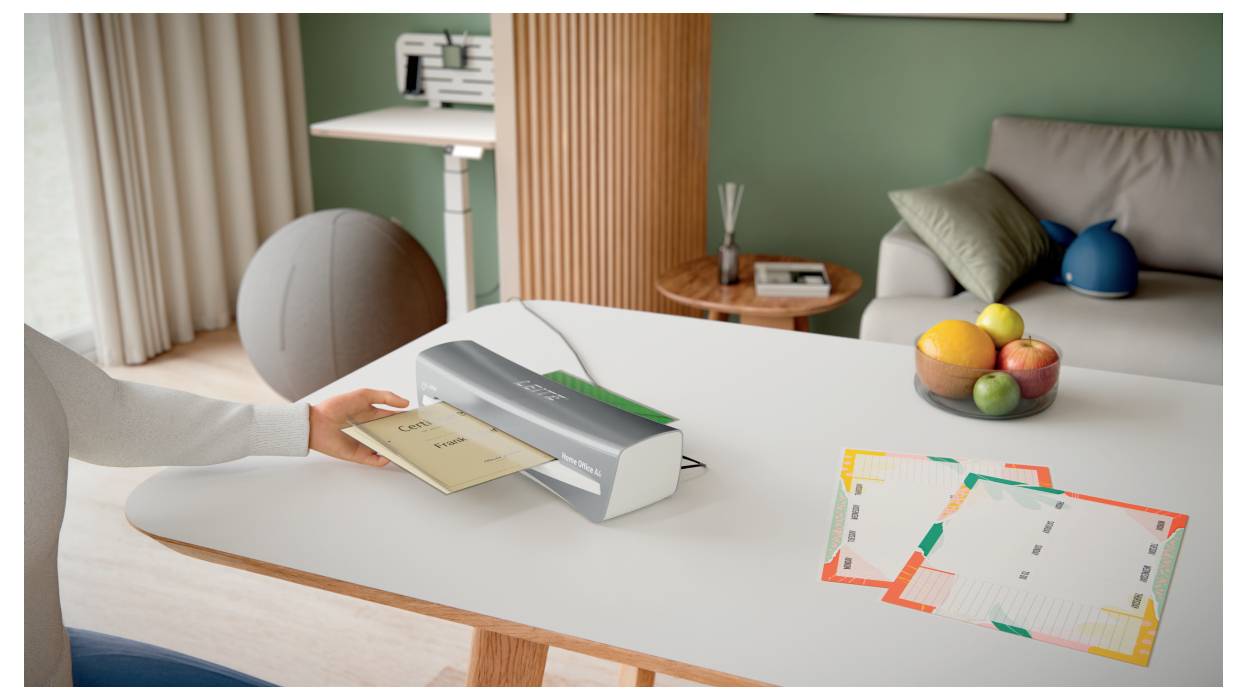 Eine Person laminiert ein Dokument in einem modernen Büro mit einem Tisch und einer Obstschale. Im Hintergrund sind Papiere, ein Regal und Sitzmöglichkeiten sichtbar.