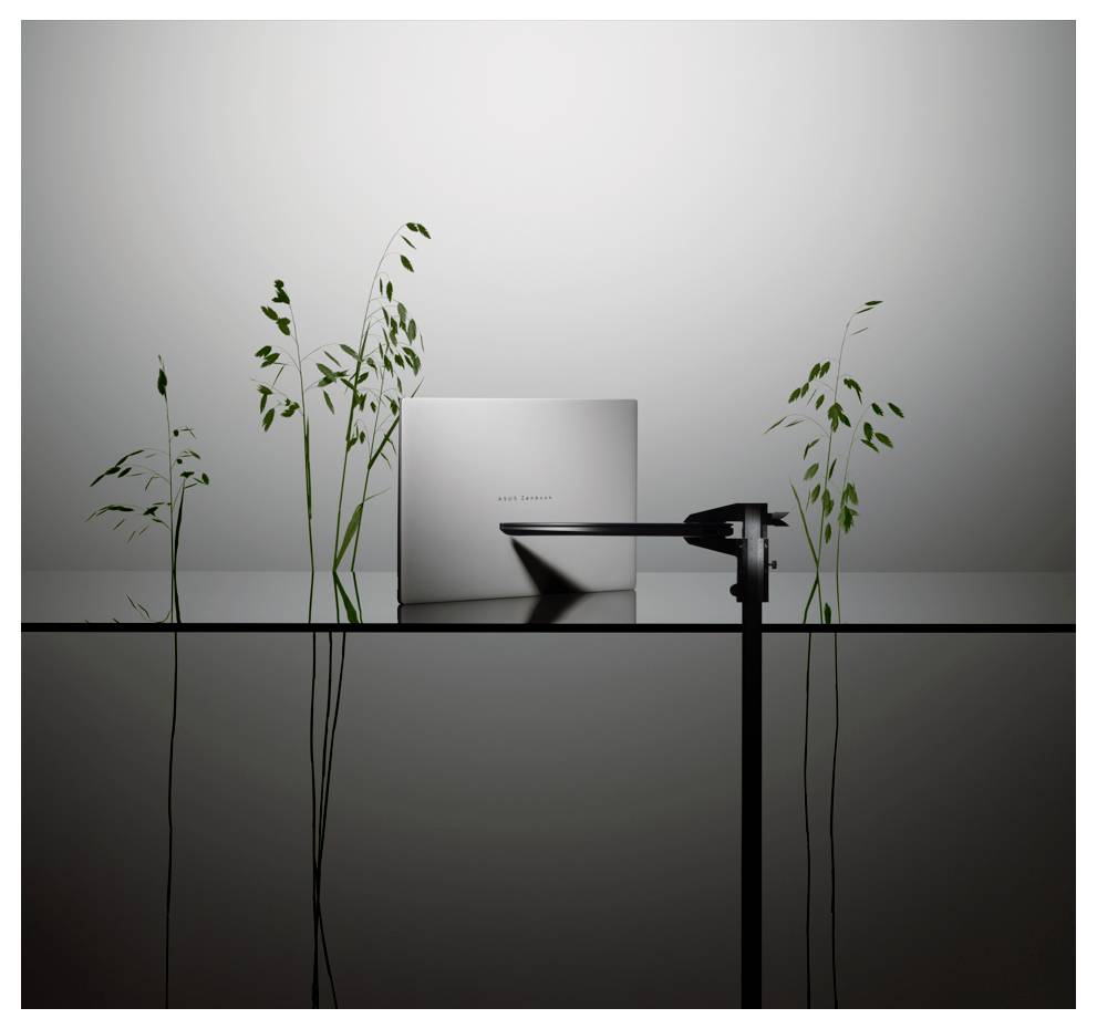Ein eleganter Laptop auf einer reflektierenden Oberfläche, umgeben von hohen Pflanzen, mit einem mechanischen Arm, der unter weicher Beleuchtung auf den Bildschirm zeigt.