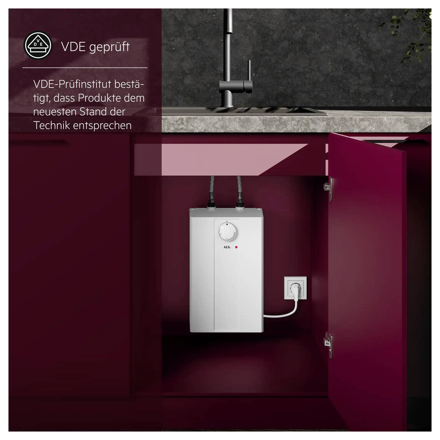 Ein weißer Warmwasserspeicher ist in einem roten Küchenschrank unter einem Waschbecken installiert. Ein Etikett zeigt an, dass er VDE-zertifiziert für technische Normen ist.
