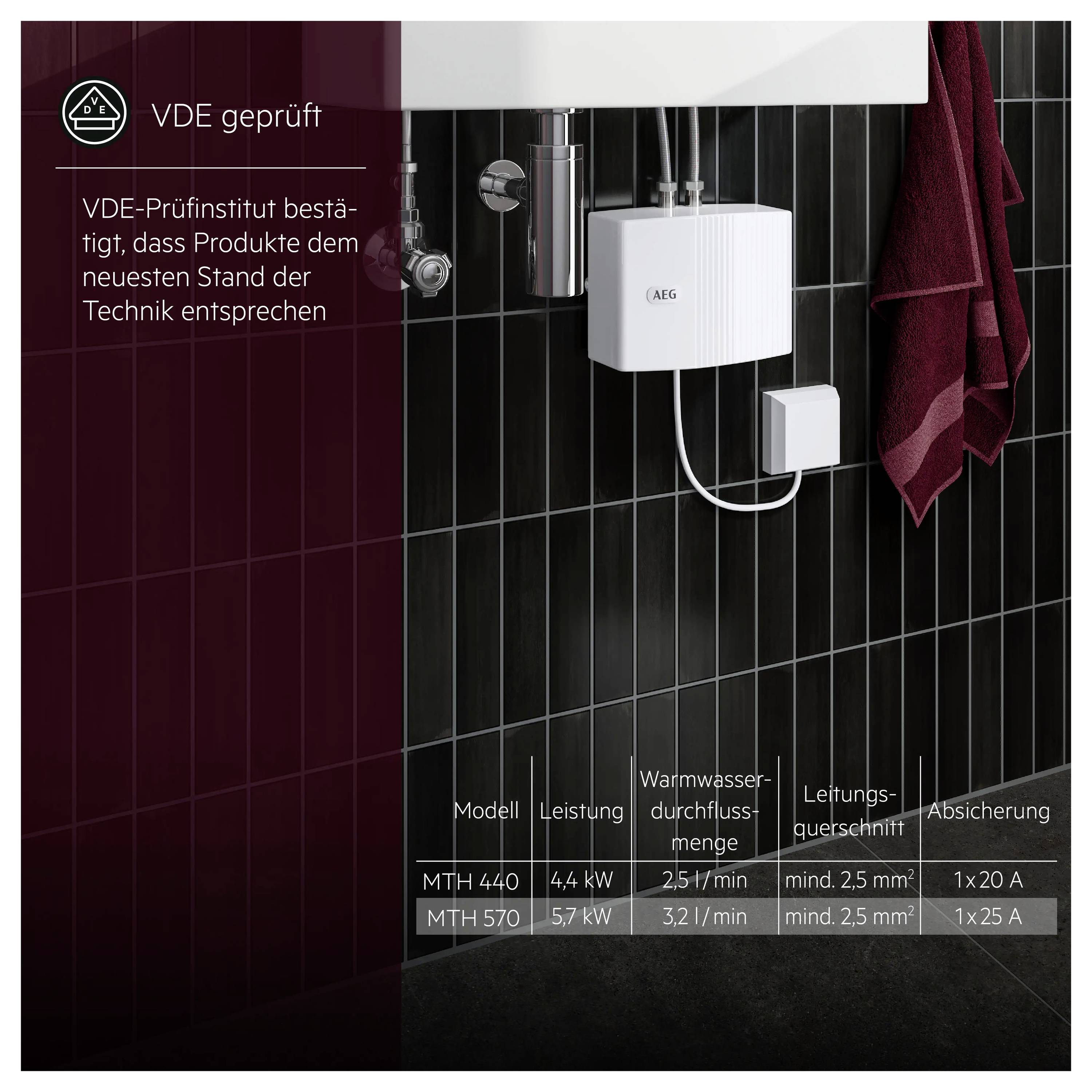 Ein kleiner weißer Warmwasserbereiter unter einem Waschbecken installiert mit freiliegenden Rohren an einer schwarzen gekachelten Wand. Ein rotes Handtuch hängt auf der rechten Seite. Technische Spezifikationen und der Text „VDE geprüft" sind sichtbar.