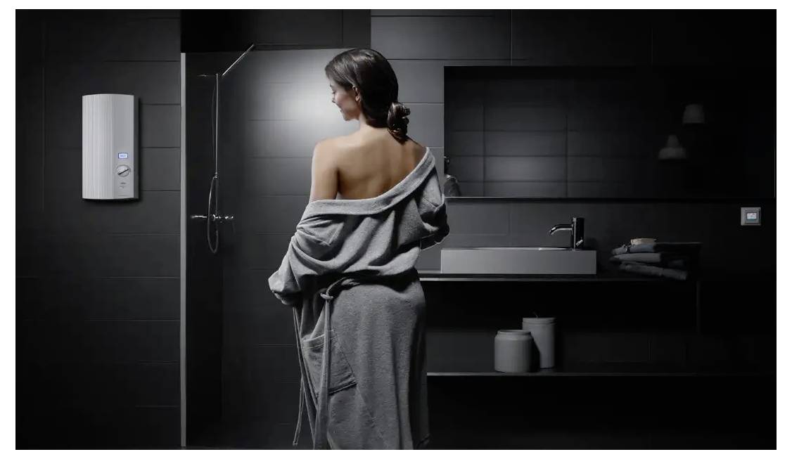 Eine Frau mit einem um sie herum drapiertem Handtuch steht in einem modernen, dunkelfliesigen Badezimmer neben einem wandmontierten Warmwasserbereiter.