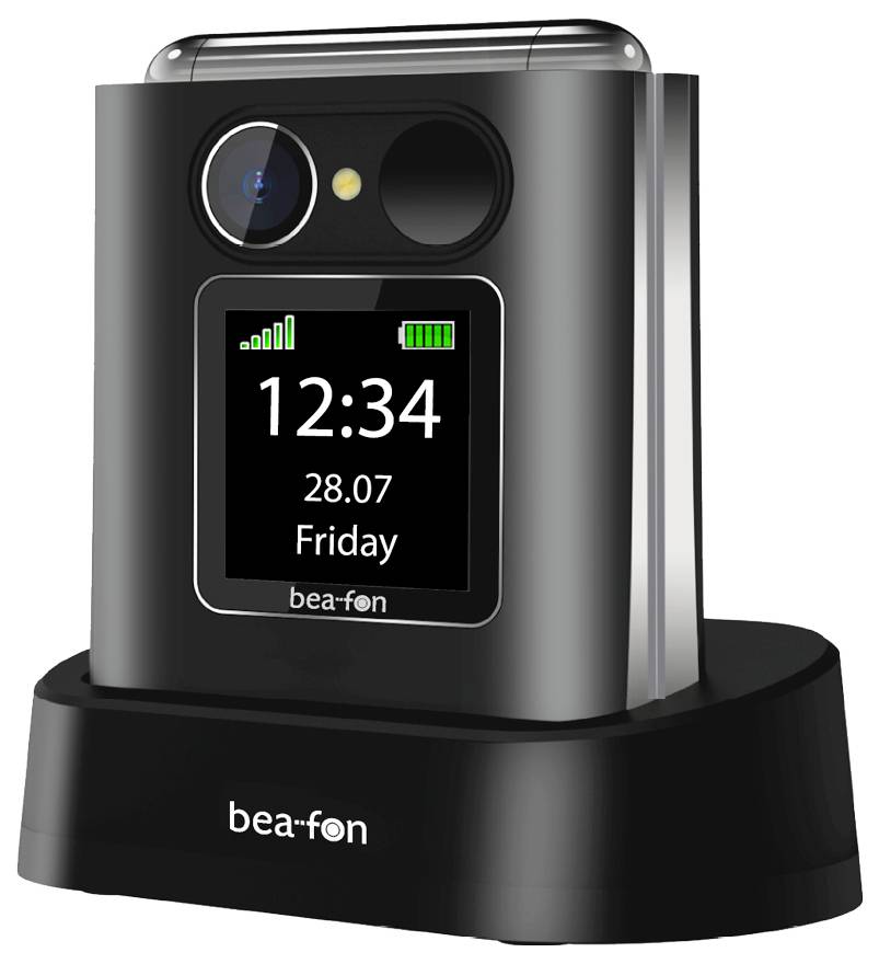 Ein schwarzes Klapptelefon auf einer Ladestation, das auf seinem Display „12:34" und „28.07 Freitag" anzeigt, mit sichtbaren Signal- und Batterieanzeigebalken.
