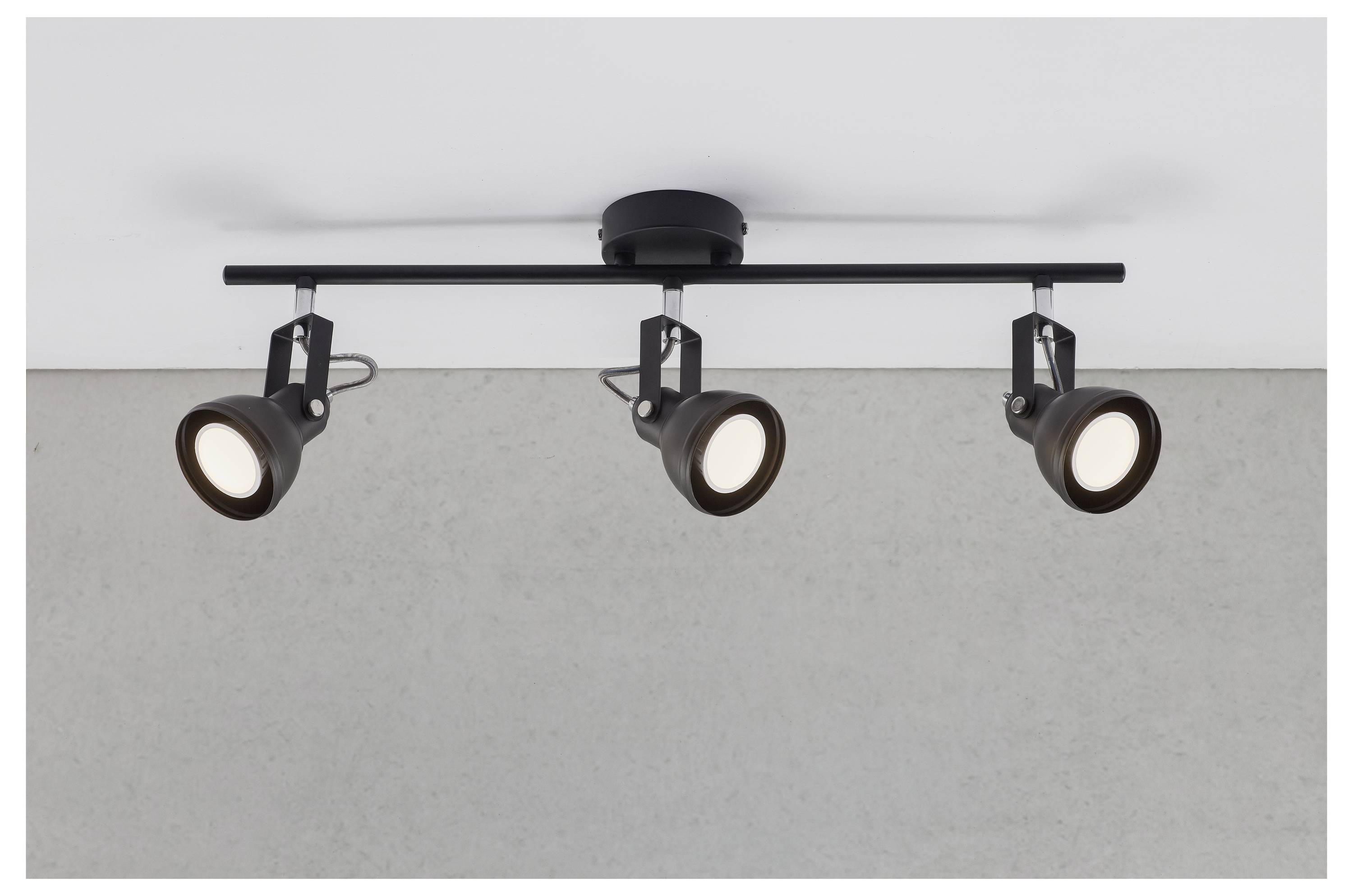Drei verstellbare Deckenspotlichter auf einer schwarzen Metallschiene, nach unten abgewinkelt, an einer weißen Decke gegen einen grauem Wandhintergrund montiert.
