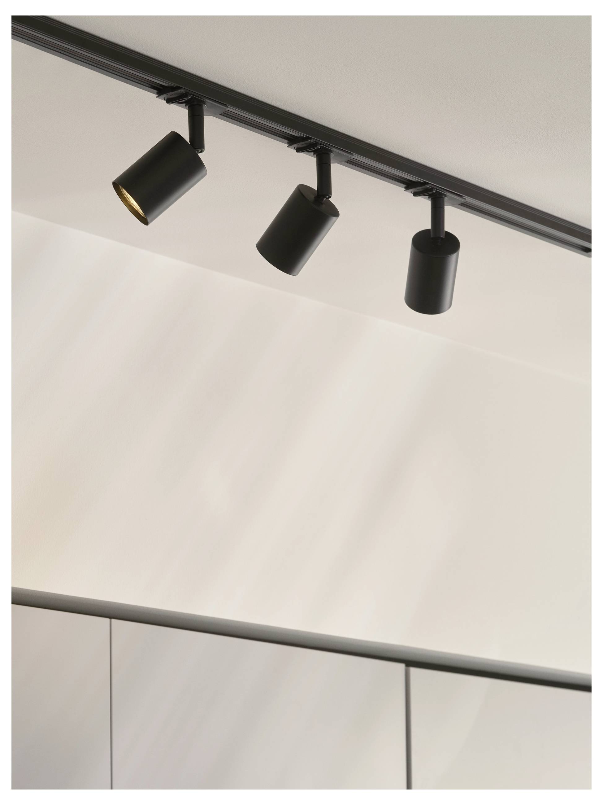 Drei schwarze Schienenstrahler an einer Decke, zum Leuchten einer Wand ausgerichtet, werfen weiches Licht auf eine neutralfarbige Oberfläche.