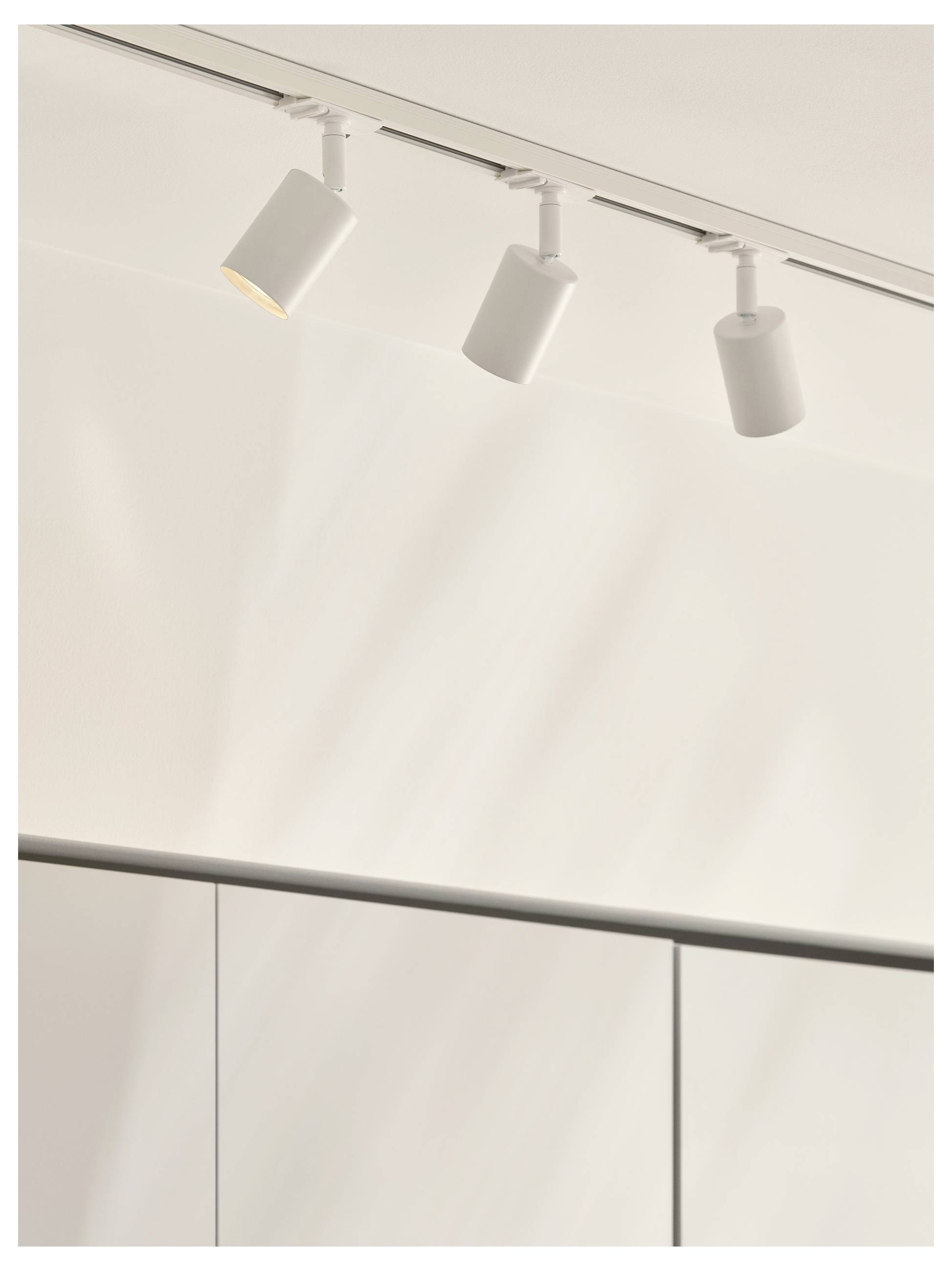 Deckenbahnleuchten mit drei weißen zylindrischen Leuchten, die einen minimalistischen Raum mit glattem, hellfarbigem Wandbelag beleuchten.