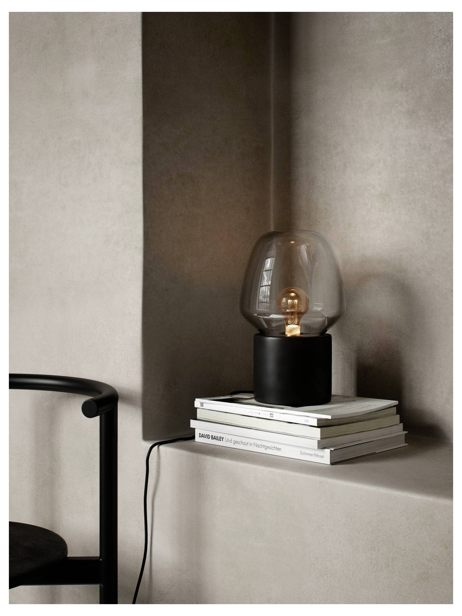 Eine moderne schwarze Tischleuchte mit einem klaren Glasschirm steht auf einem Stapel Bücher vor einer neutral getönten Wand. Die Lampe ist eingeschaltet.