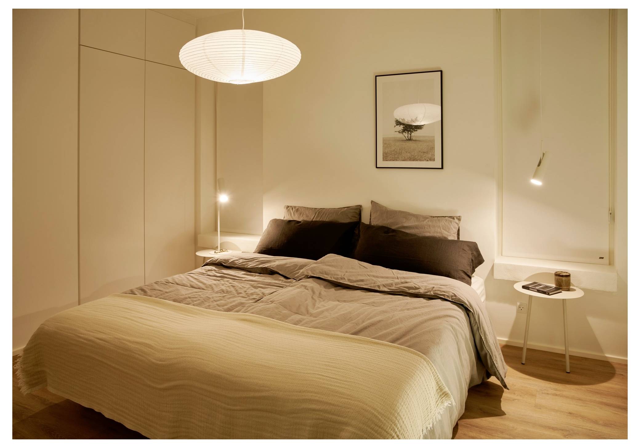 Ein gemütliches Schlafzimmer mit einem ordentlich gemachten Bett, sanfter Beleuchtung und minimalistischer Einrichtung. Ein Nachttisch hält ein Buch, und ein Kunstwerk hängt über dem Bett.