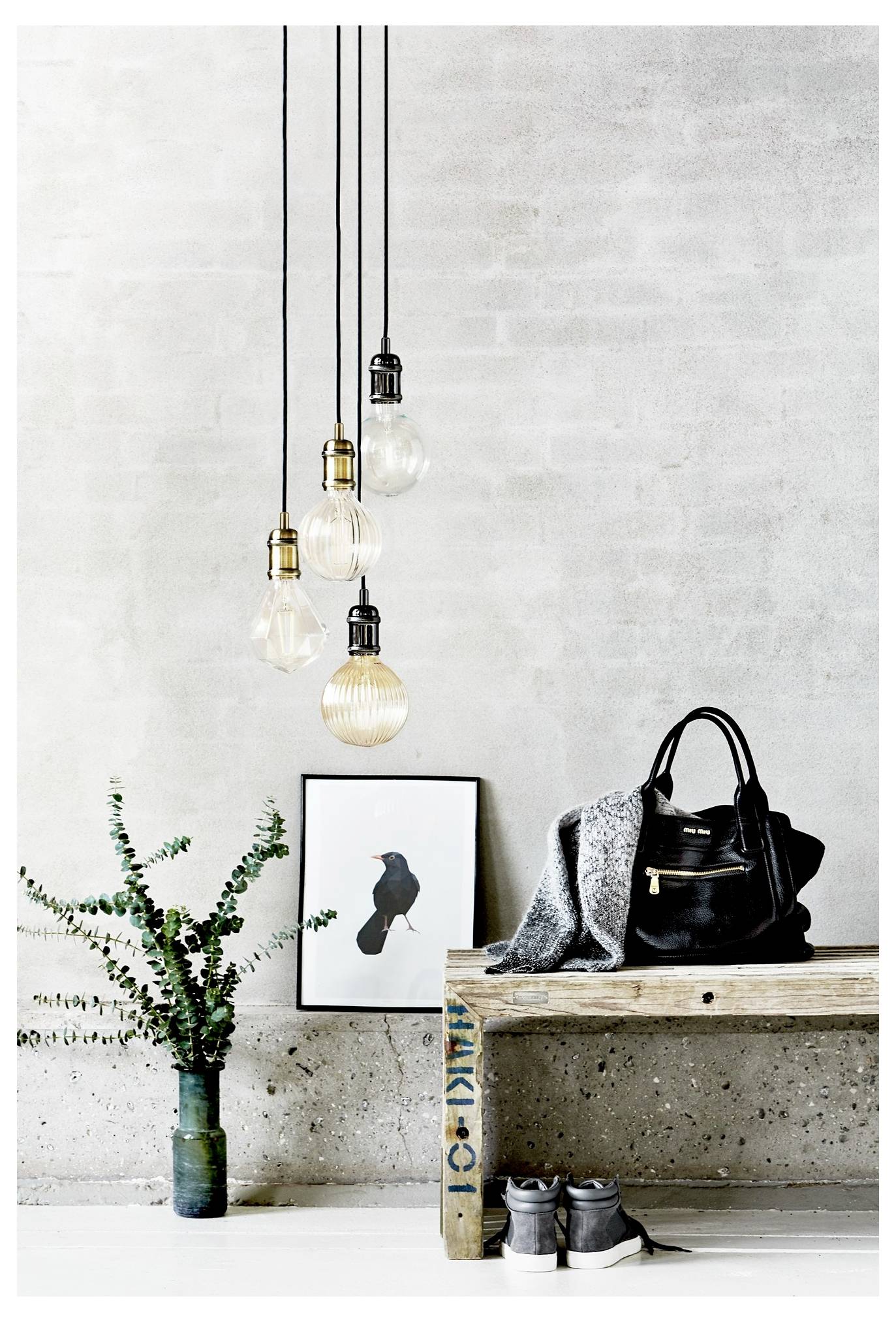 Ein minimalistisches Interieur zeigt eine hängende Gruppe von vier dekorativen Glühbirnen, eine hölzerne Bank mit einer schwarzen Tasche und einem grauen Pullover, Sneaker darunter, einen gerahmten Vogelprint und eine Vase mit Grün.