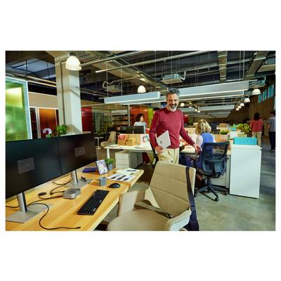 Mann, der lächelnd einen Laptop in einem modernen Büro hält; Mitarbeiter arbeiten an Schreibtischen mit Computern im Hintergrund; lebendige und kollaborative Atmosphäre.
