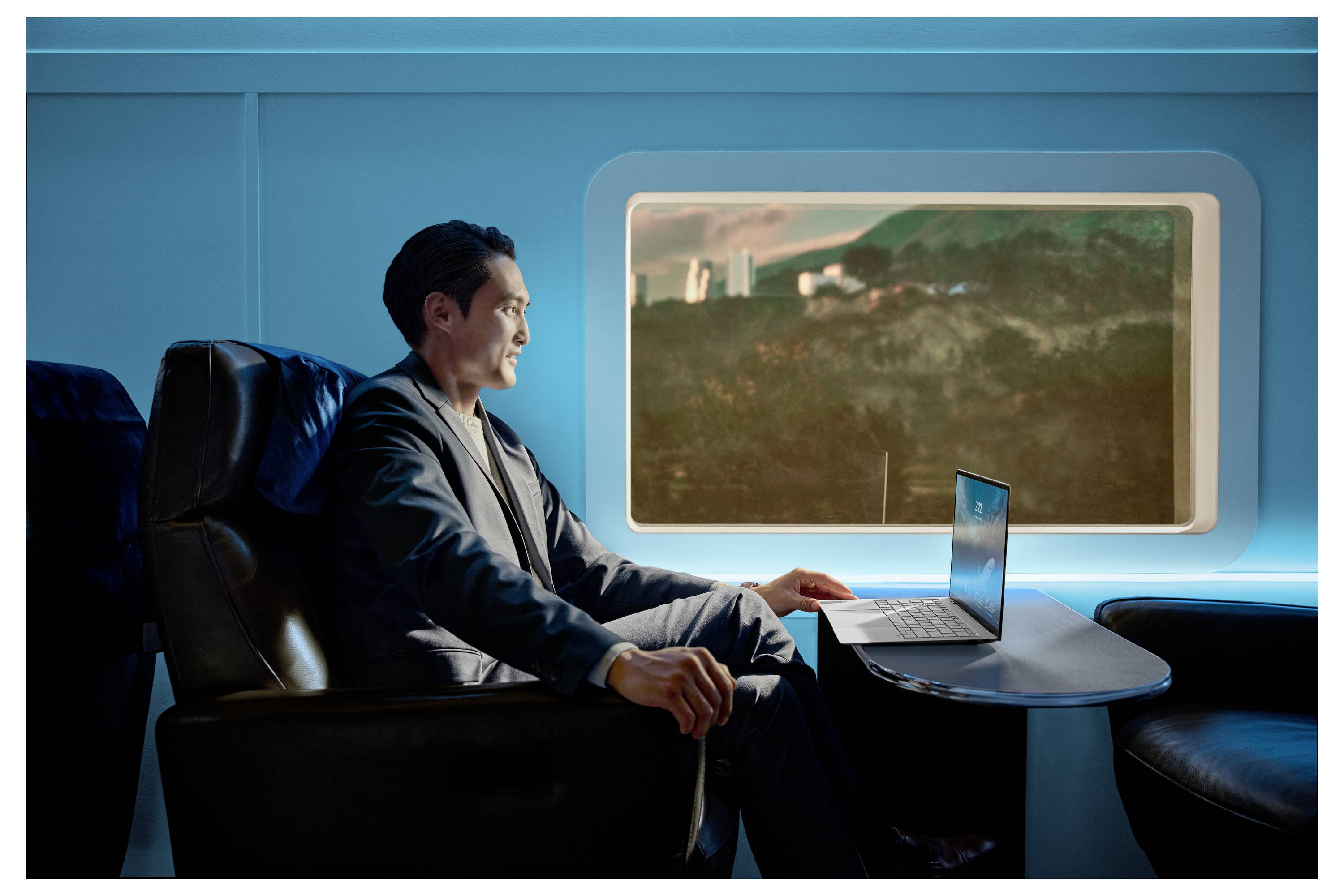 Person sitzt in einer futuristischen Zugkabine, nutzt einen Laptop auf einem Tisch und hat durch ein großes Fenster einen Blick auf eine Landschaft.