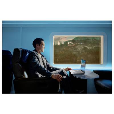 Person sitzt in einer futuristischen Zugkabine, nutzt einen Laptop auf einem Tisch und hat durch ein großes Fenster einen Blick auf eine Landschaft.