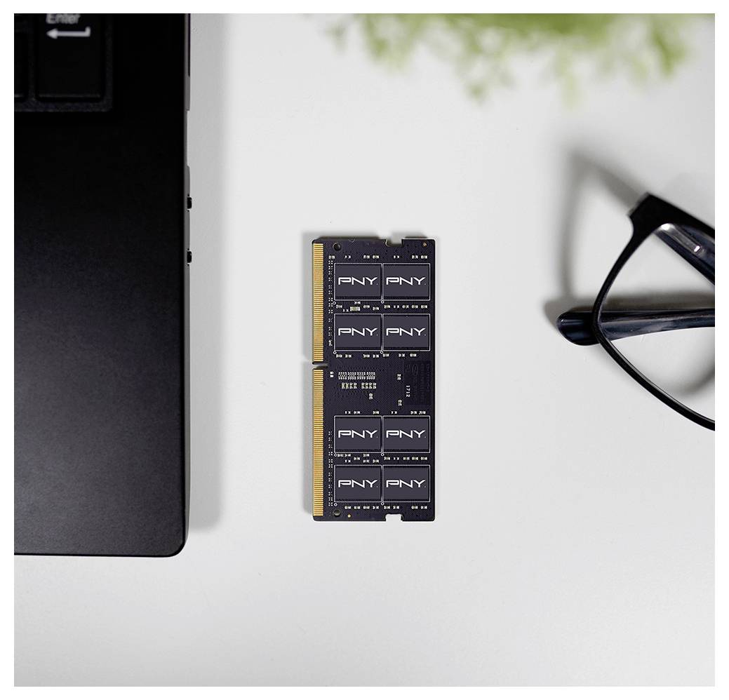 Ein PNY-RAM-Modul liegt auf einem weißen Schreibtisch neben einer Laptopecke und einer Brille, was auf eine Tech-Ausstattung oder eine Arbeitsumgebung hindeutet.
