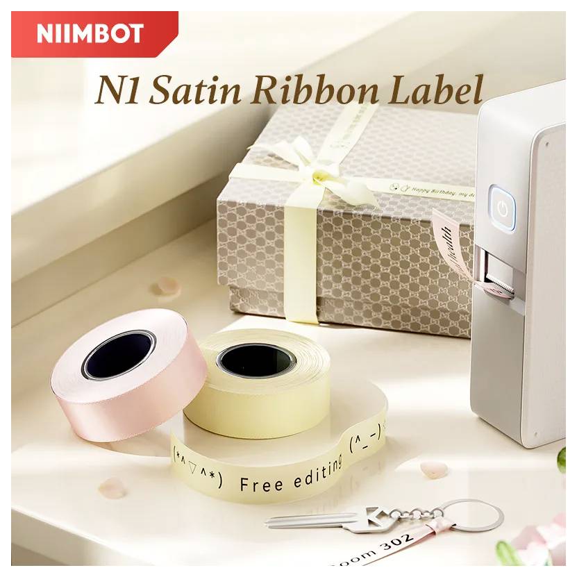 NIIMBOT N1 Satinband-Etikettendrucker mit rosa, gelben und cremefarbenen Etikettenballen neben einer Geschenkbox und einem Schlüsselanhänger auf einer Tischplatte.