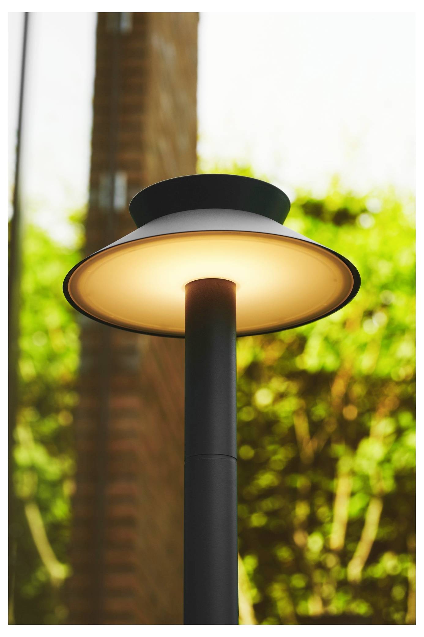 Eine moderne Straßenlampe, die tagsüber beleuchtet ist, vor einem unscharfen Hintergrund aus Grün und einer Backsteinmauer.