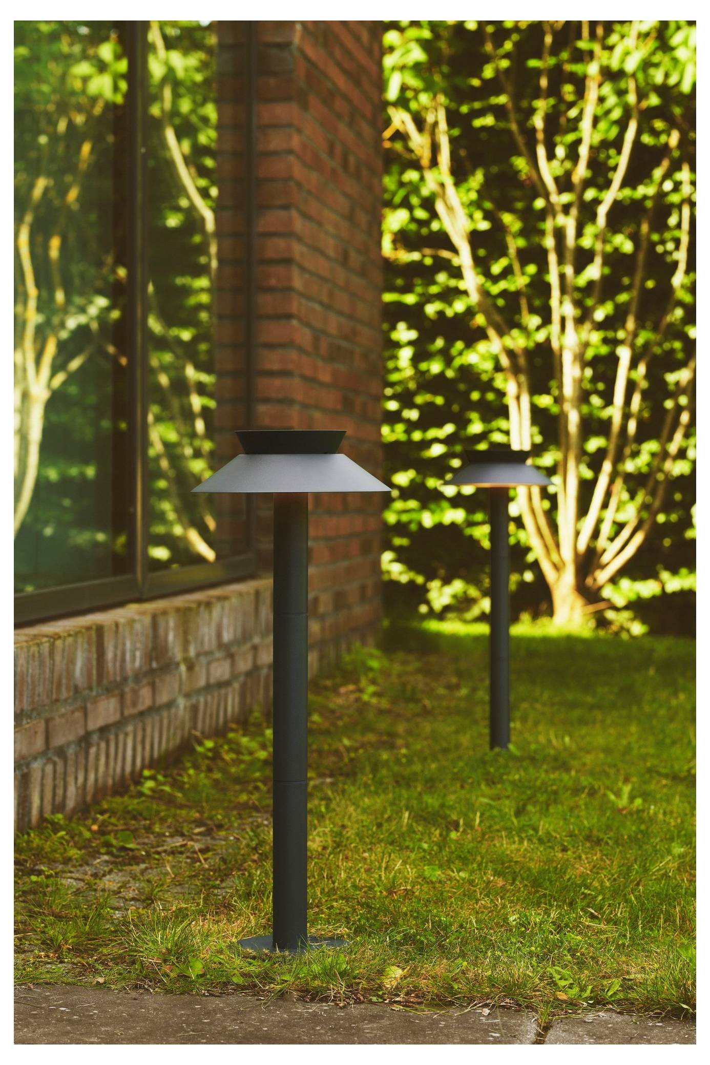 Zwei Außen-Solargartenleuchten stehen auf einem Rasen neben einem Backsteingebäude mit einem Glasfenster, umgeben von grünem Laub.
