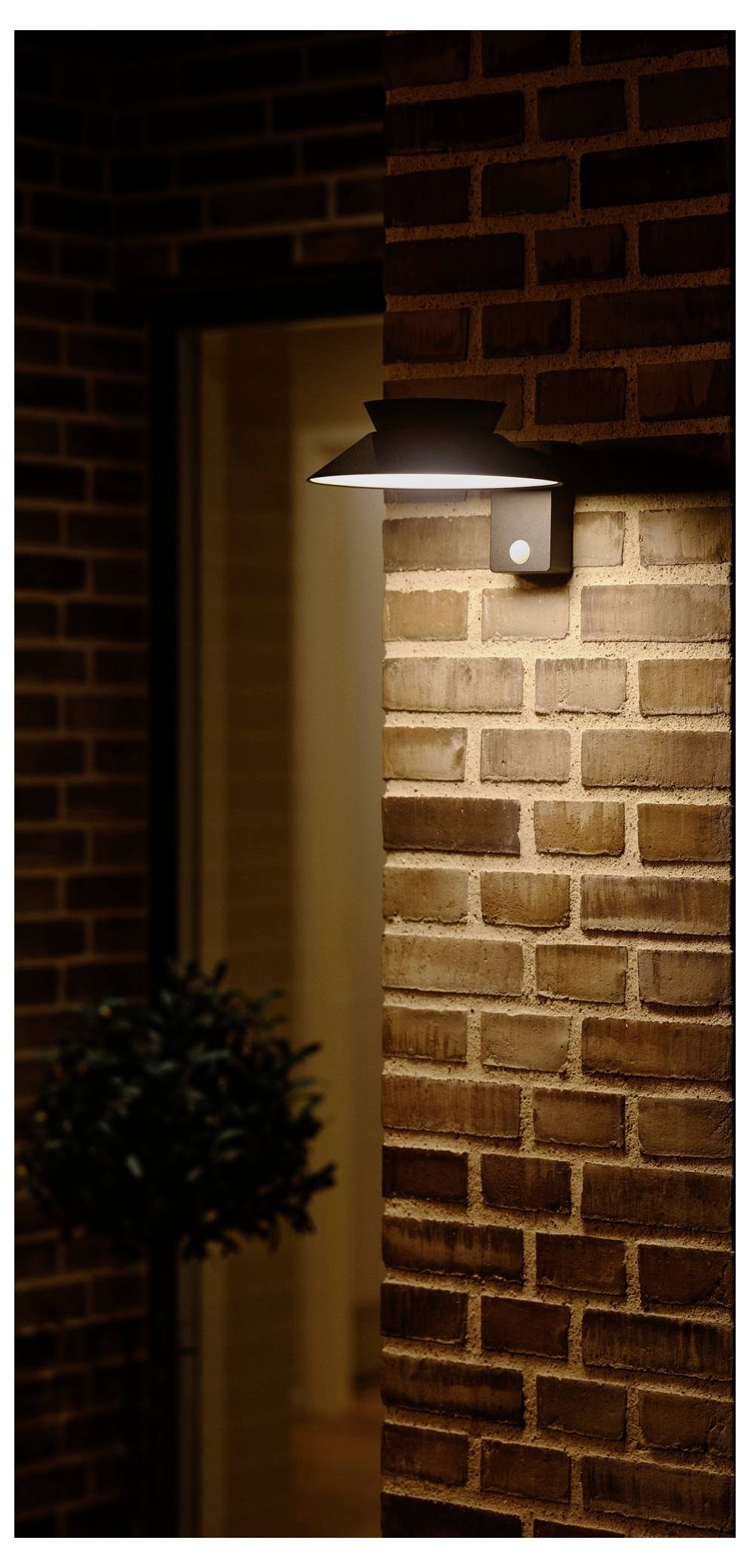 Ein modernes Außenwandlicht beleuchtet eine Backsteinmauer außerhalb eines Gebäudes und schafft eine warme Atmosphäre. Eine kleine Topfpflanze befindet sich in der Nähe.