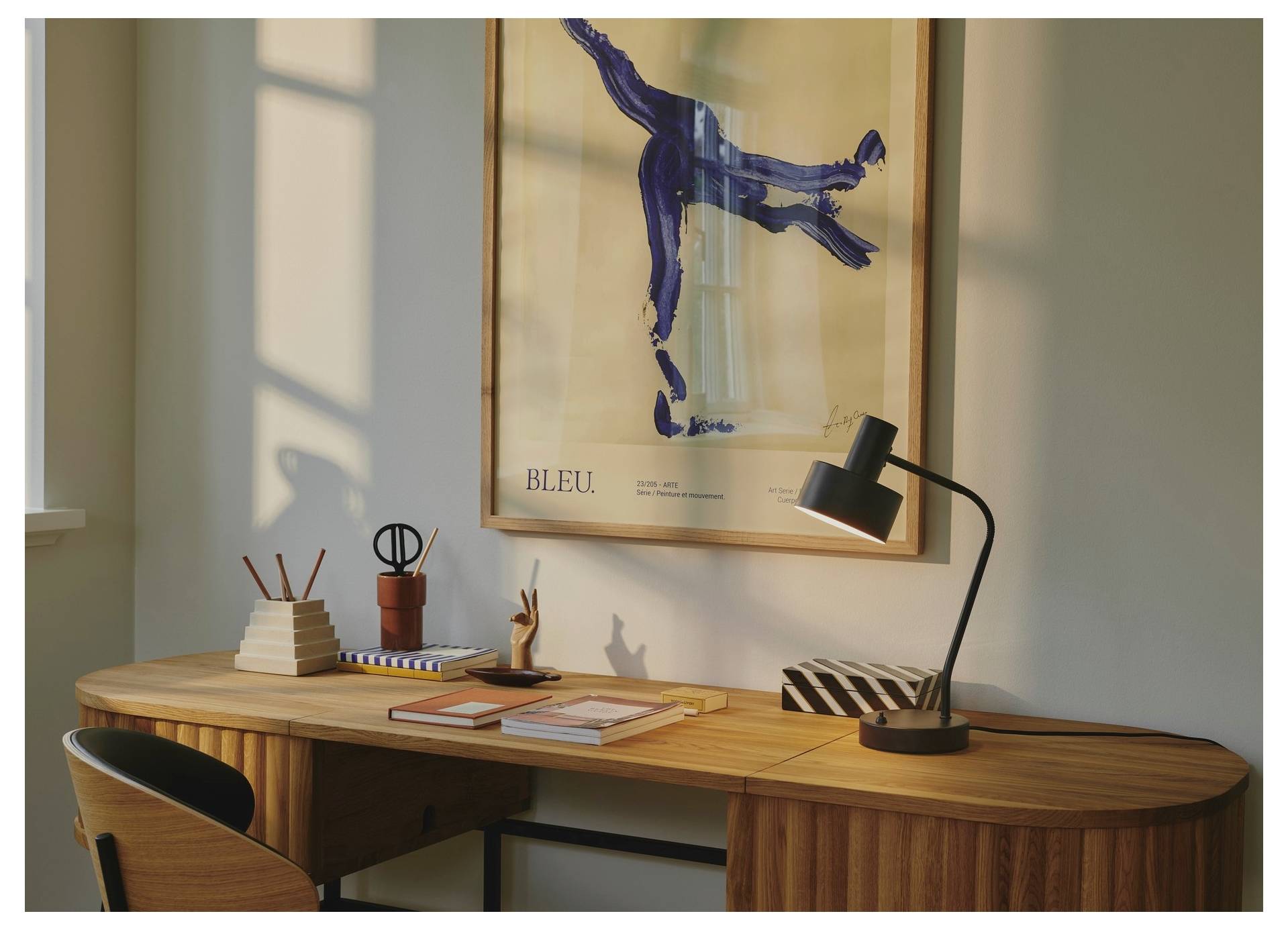 Ein Holzschreibtisch mit Büchern, Notizblöcken und einer Lampe in einem warm beleuchteten Raum. Ein Poster mit dem Titel „BLEU" und abstraktem Kunstwerk hängt an der Wand.