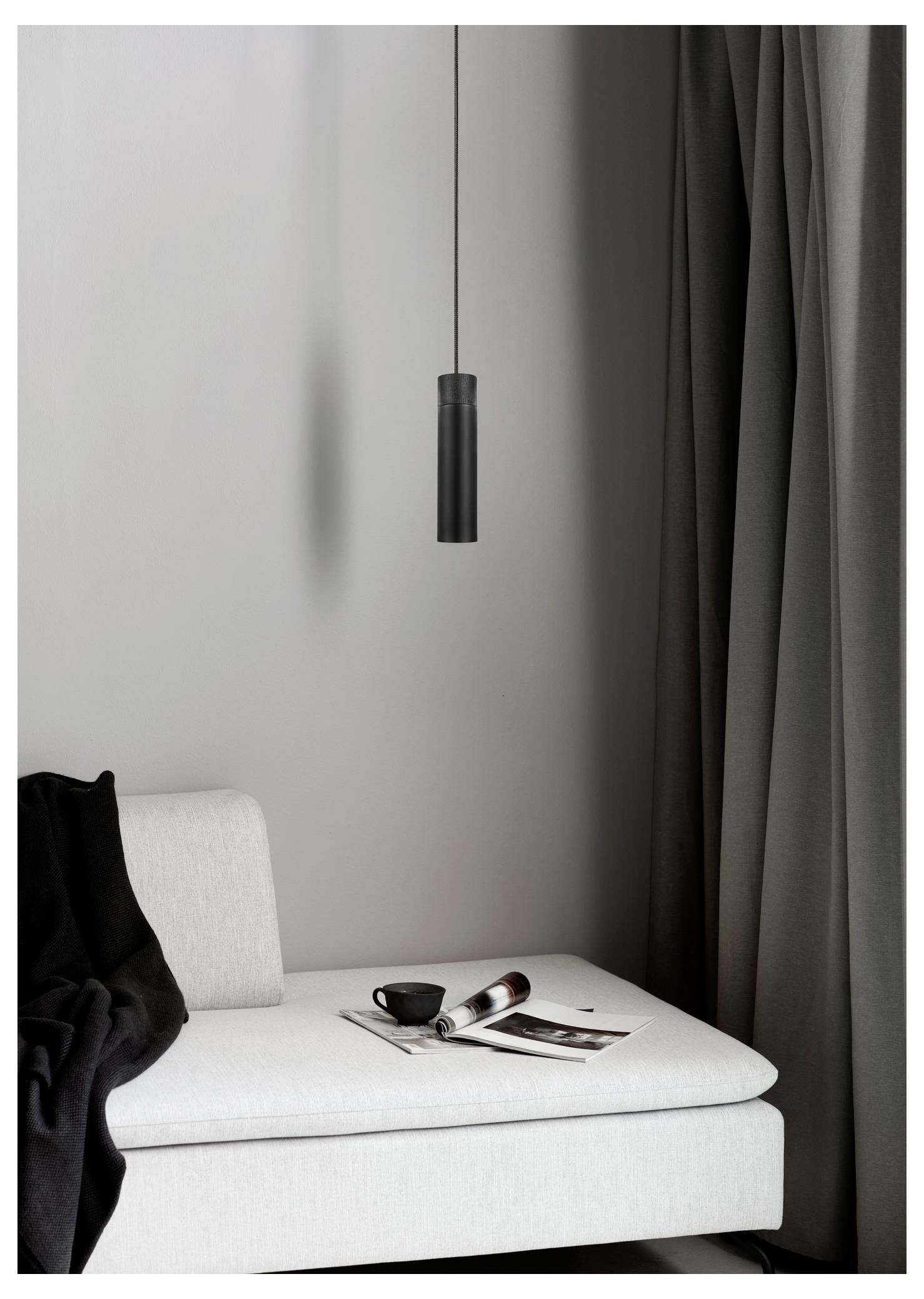 Ein minimalistisches Zimmer mit einer weißen Chaiselongue, einer schwarzen Wurfdecke, einem aufgeschlagenen Magazin und einer Kaffeetasse. Eine Pendelleuchte hängt darüber.