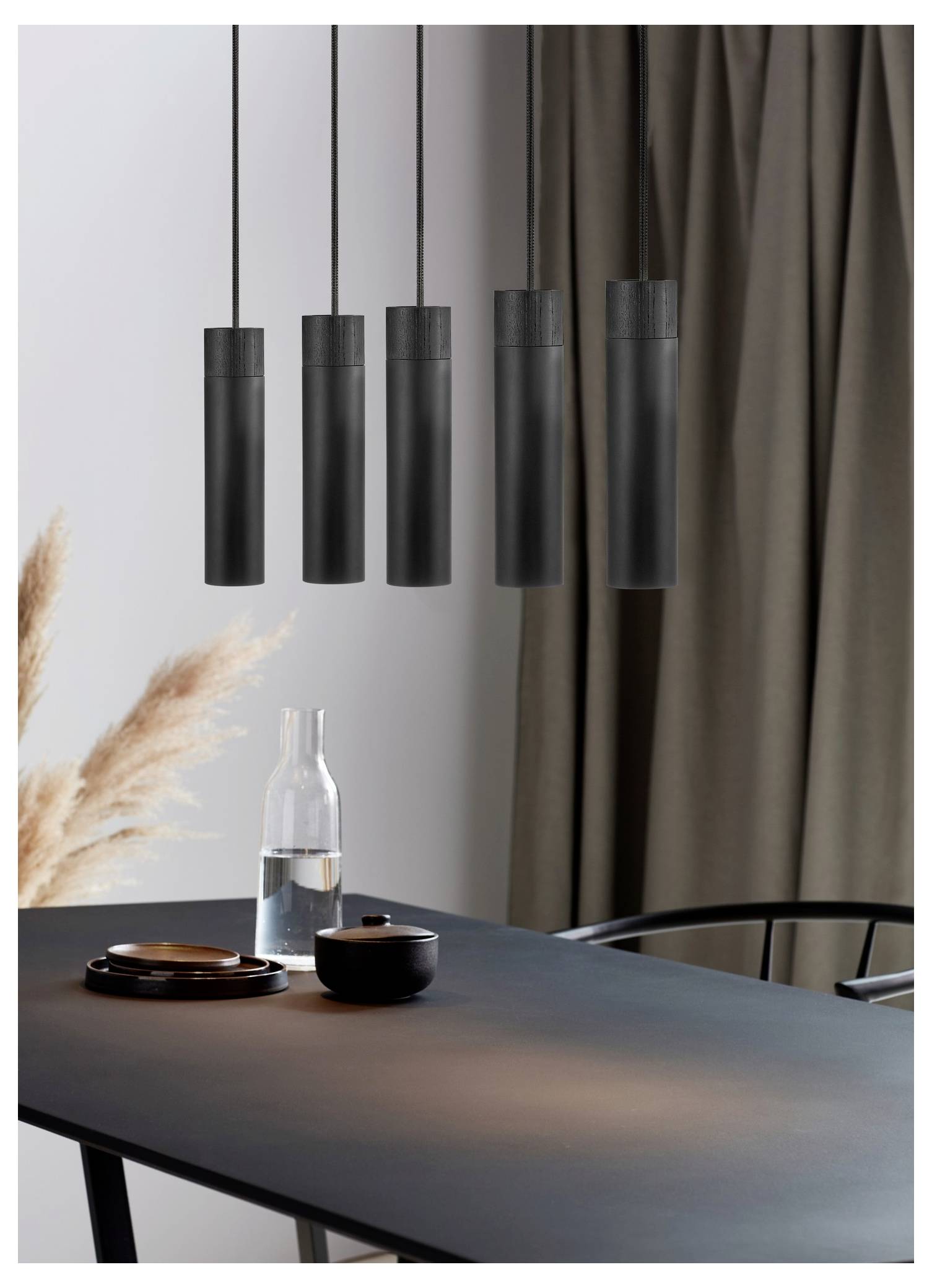Ein moderner Essbereich verfügt über einen eleganten schwarzen Tisch mit einer Wasserflasche, einer Tasse und fünf Pendelleuchten darüber. Getrocknetes Pampasgras schmückt den Raum.