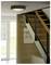 Ein modernes Interieur mit einer Holztreppe, schwarzem Geländer und einer runden Deckenleuchte. Ein Poster hängt an der Wand neben großen Fenstern.