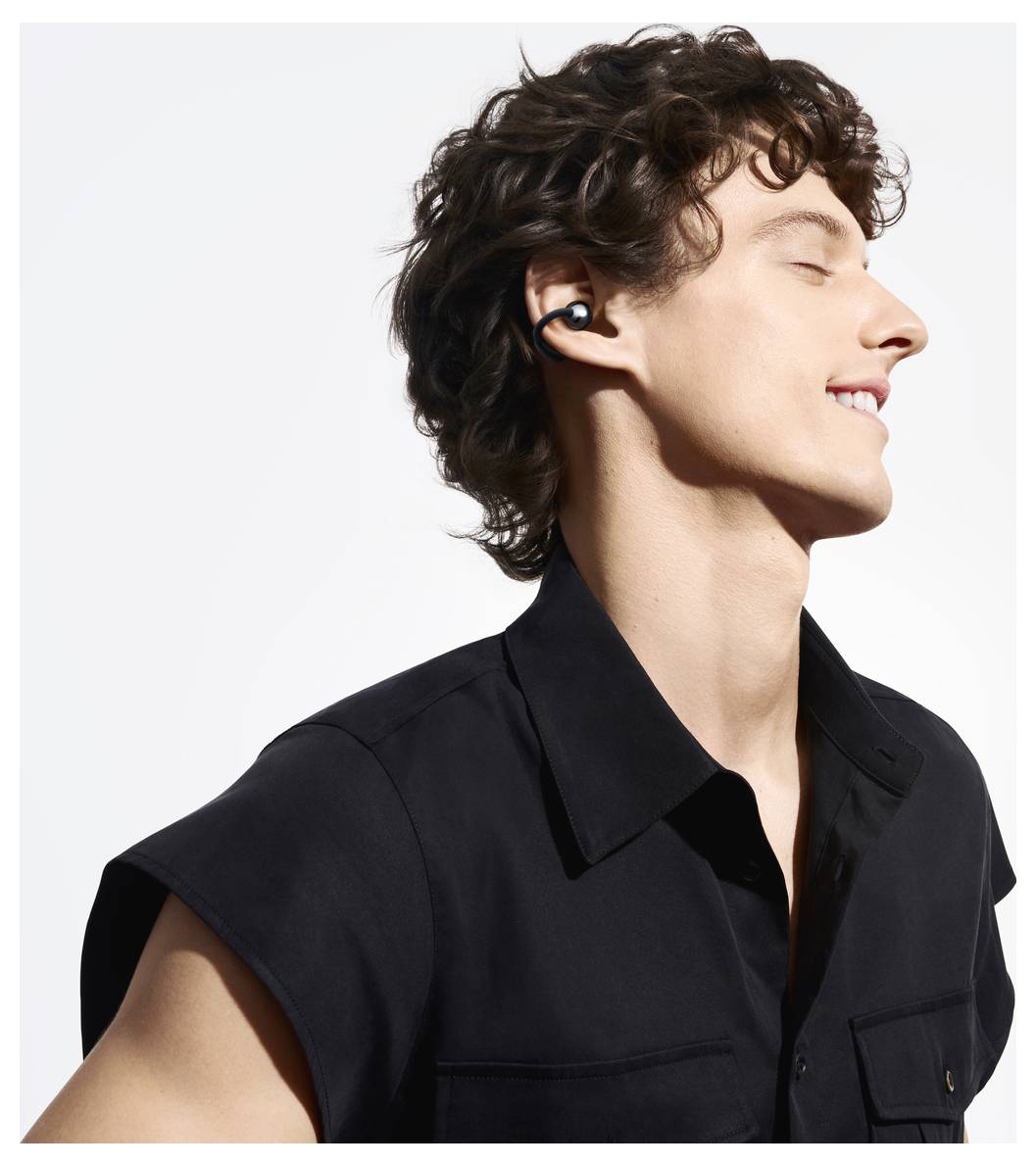 Person mit lockigem Haar, trägt kabellose Ohrhörer und ein schwarzes Hemd mit Kragen, lächelt mit geschlossenen Augen vor einem hellen Hintergrund.