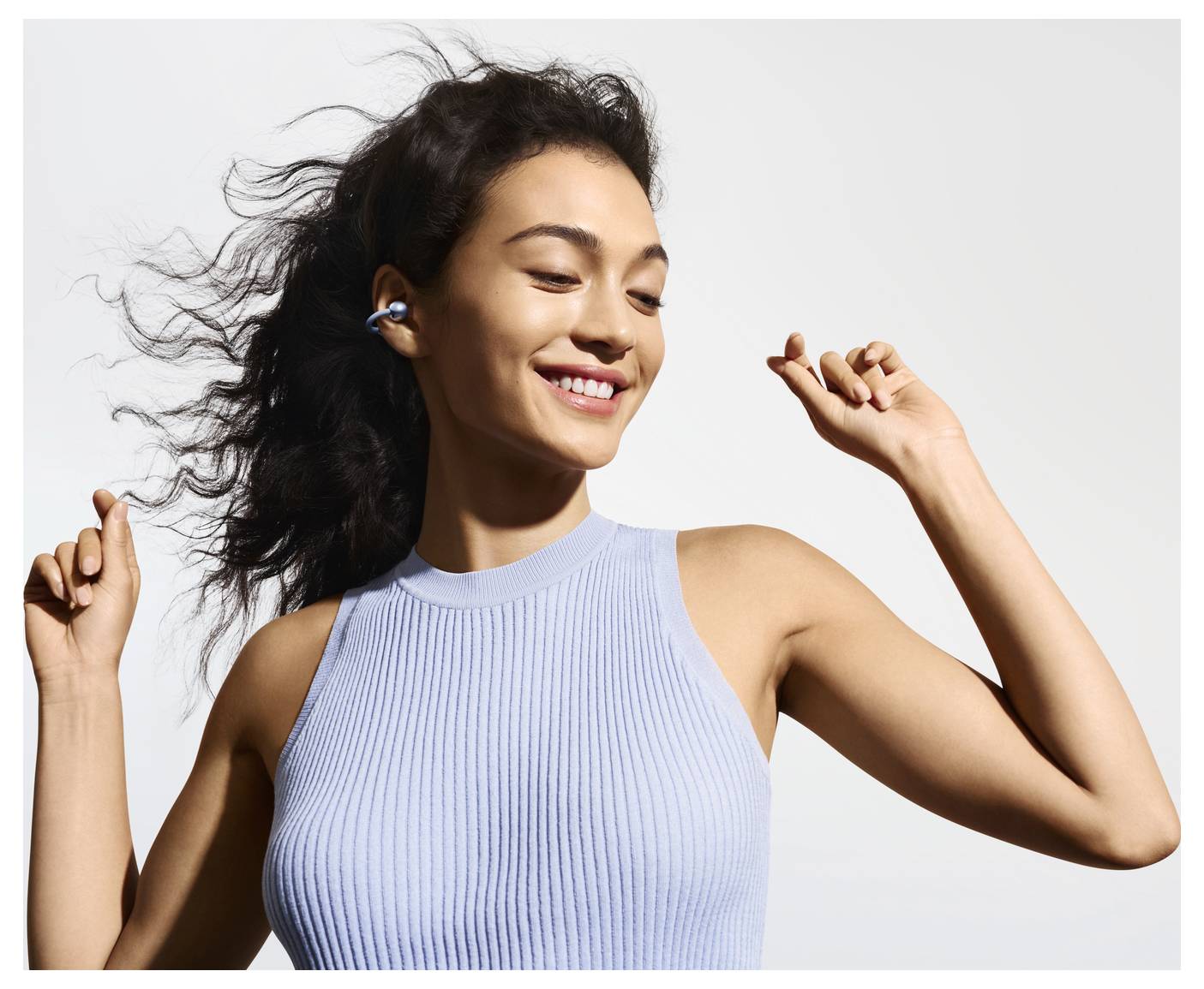 Eine Frau mit dunklem, welligem Haar tanzt freudig, während sie kabellose Ohrhörer trägt, gekleidet in ein ärmelloses, blaues geripptes Oberteil, lächelnd mit geschlossenen Augen.