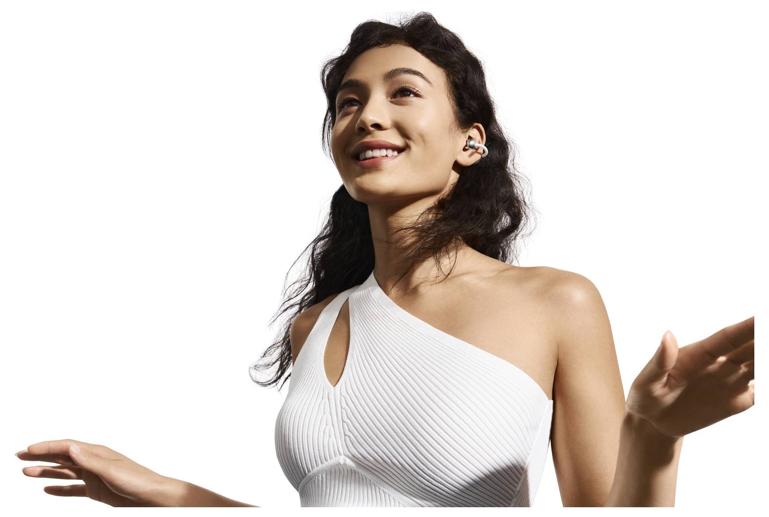 Eine Frau lächelt, während sie kabellose Ohrhörer trägt, steht vor weißem Hintergrund und trägt ein weißes Oberteil mit einer Schulter.